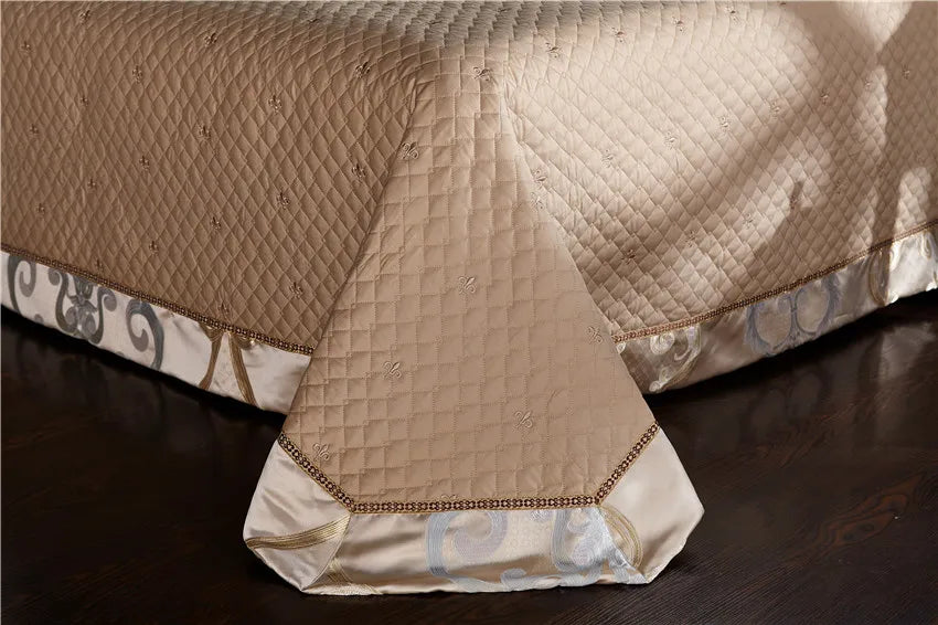 A close-up of a quilted bedspread in a champagne gold tone featuring delicate embroidery and diamond stitching. The hem is adorned with a satin border showcasing intricate baroque patterns in soft hues, adding a refined touch. The edges are accented with golden braided trim, providing an opulent finish that enhances the luxurious design.

