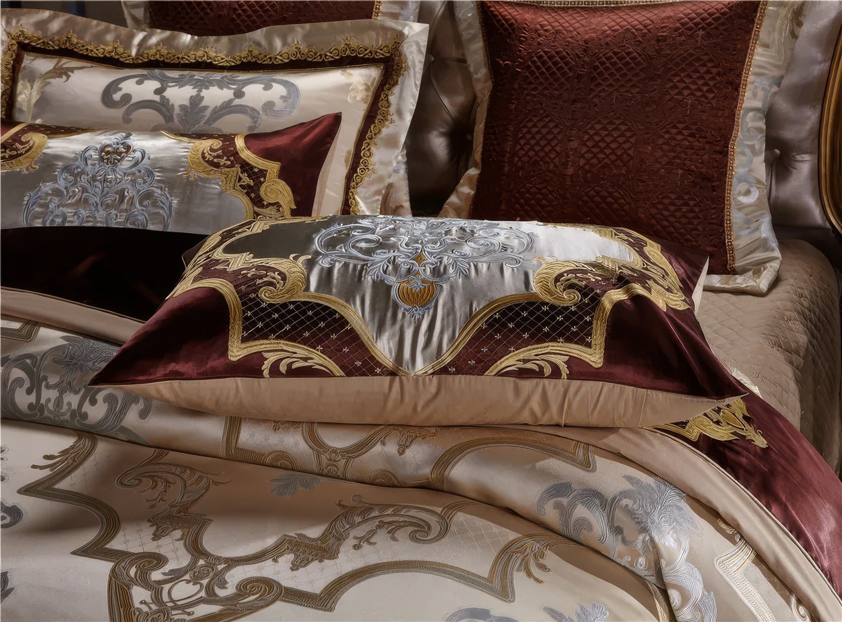 A close-up view of an opulent bedding set featuring luxurious burgundy and gold accents. The pillowcases are adorned with intricate embroidery, combining baroque-inspired patterns with shimmering silver and gold threads. The rich burgundy contrasts beautifully with the champagne satin base, highlighted by quilted details and ornate borders. This elegant design adds a regal touch to any bedroom décor.

