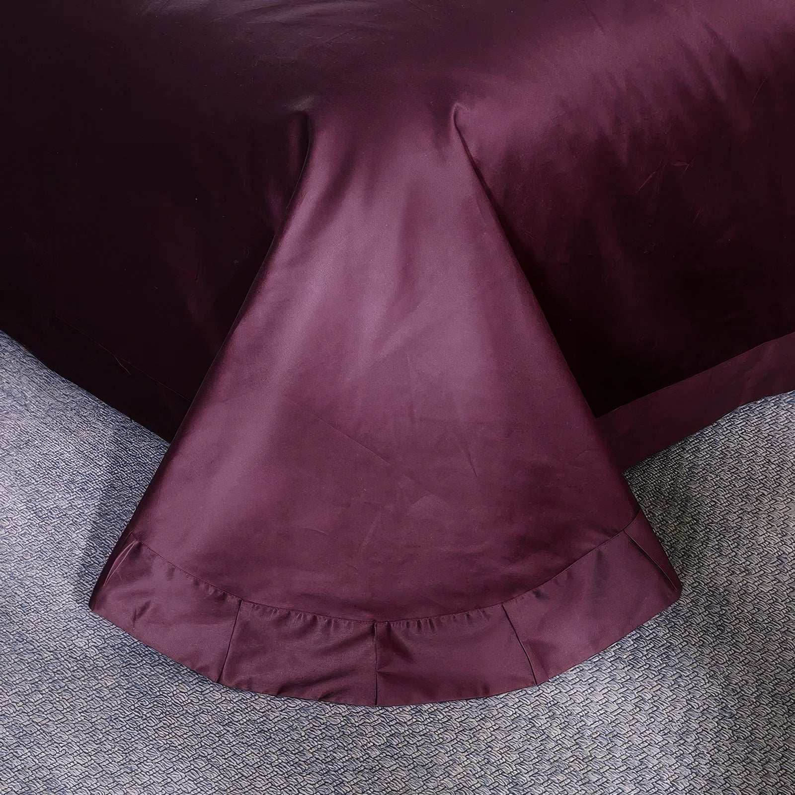 Detailed view of a maroon bed skirt, showcasing its smooth, satin-like texture and elegantly draped pleated hem, resting on a textured, neutral-toned carpet for a refined and sophisticated finish.

