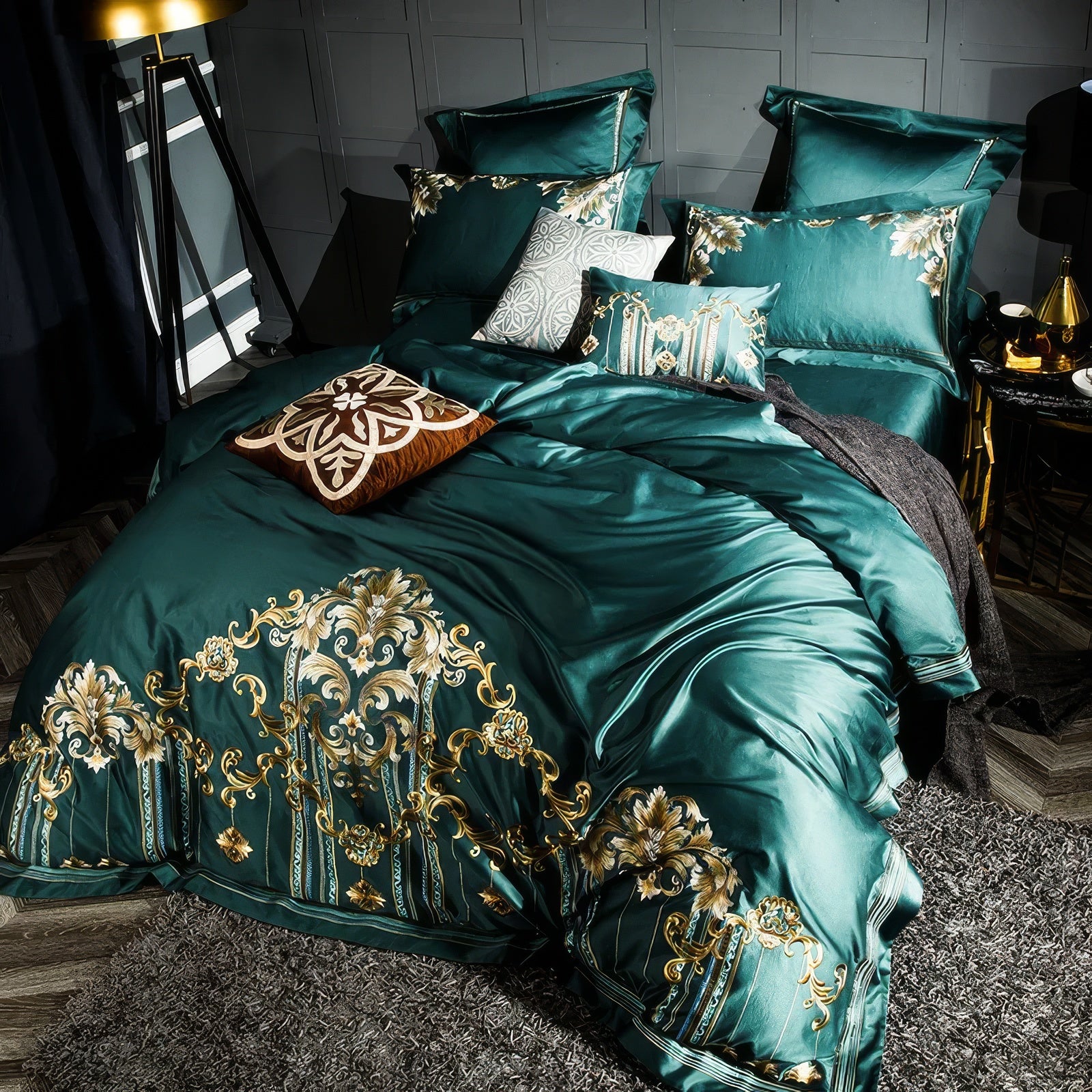 This bedding set showcases an opulent emerald green fabric, embellished with detailed golden embroidery featuring floral and ornamental motifs. The mix of accent pillows, including intricate patterns and rich textures, complements the regal design. The setting is accentuated by warm lighting and dark gray wall paneling, creating a luxurious and inviting ambiance.

