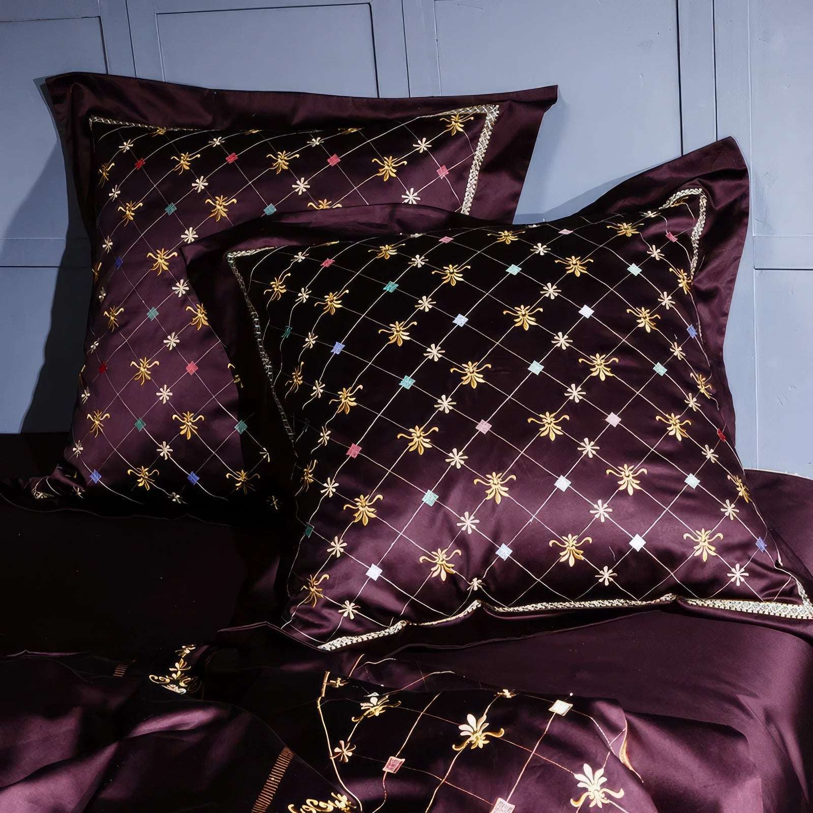 Close-up view of maroon pillowcases adorned with a crisscross pattern of fine golden threads and multicolored embroidered accents, featuring delicate floral and diamond-shaped motifs for a luxurious, detailed look.

