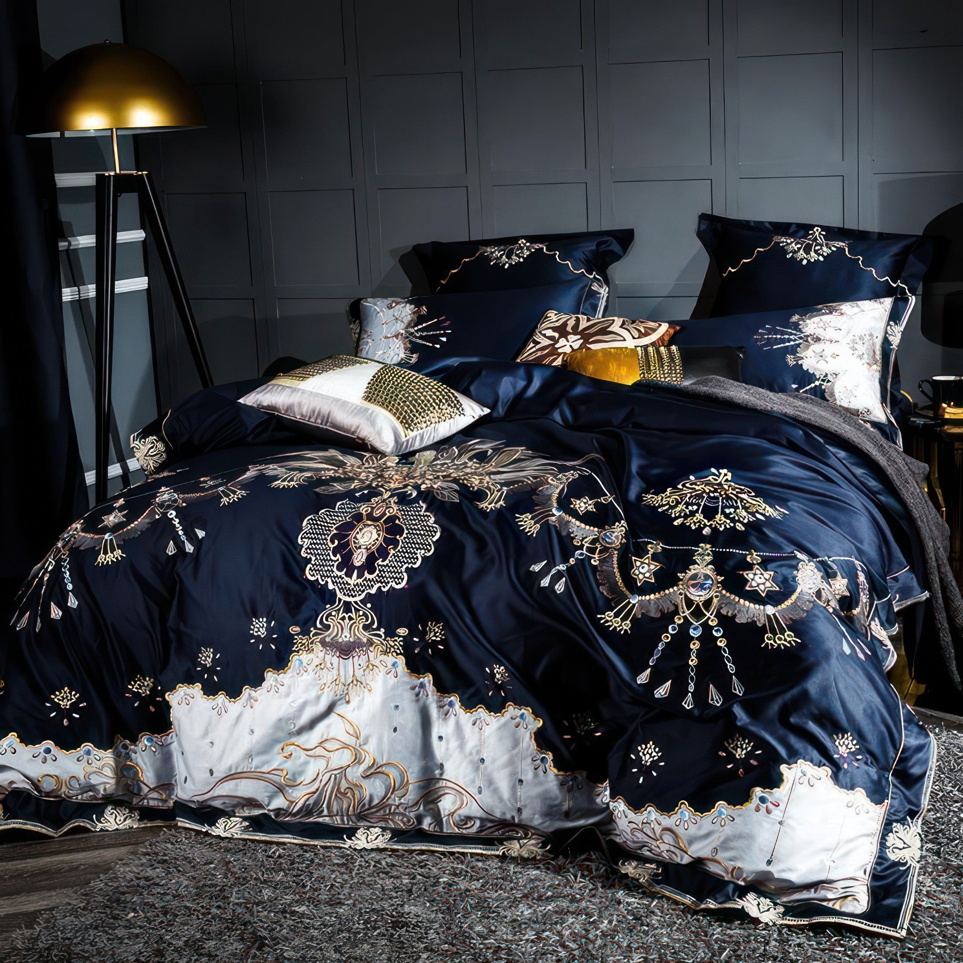 A navy and silver luxury bedding set adorned with detailed gold and white embroidery featuring floral and ornamental designs. The set includes decorative pillows in gold, white, and brown patterns, paired with a soft gray throw draped over the bed. The background features a paneled wall, complemented by a modern tripod lamp with a golden dome and a textured gray rug, creating a sophisticated and opulent bedroom ambiance.

