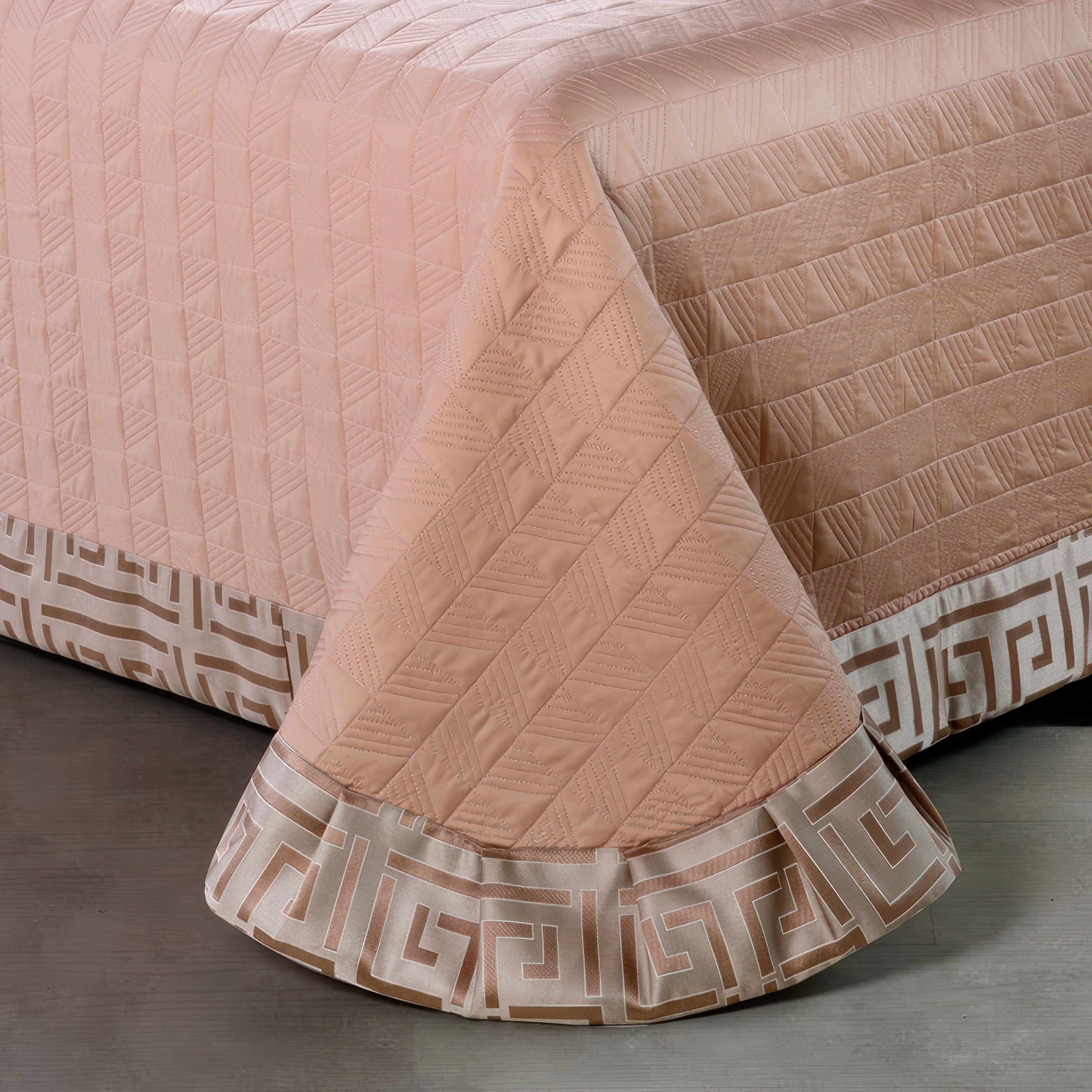 Close-up of a beige quilted bedspread featuring geometric stitched patterns and a decorative border with a metallic gold Greek key motif. The bedspread drapes gracefully over the edge, highlighting the intricate design details.