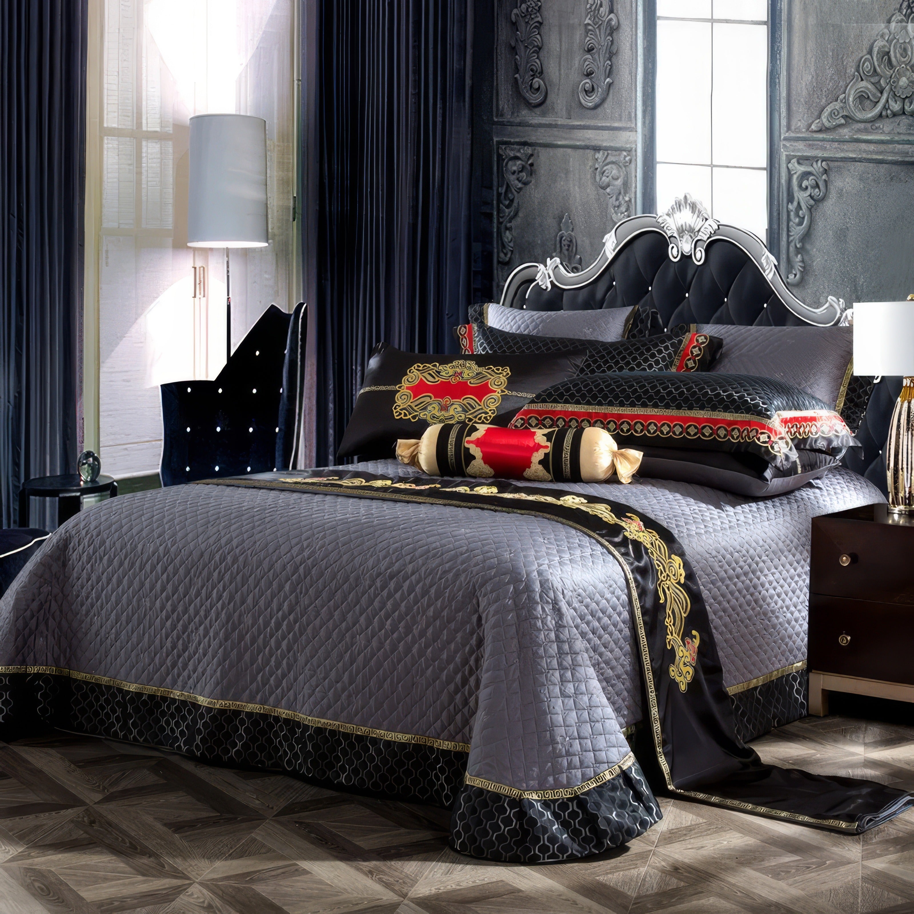 A lavishly decorated bedroom featuring a quilted gray bedspread with intricate black and gold embroidery accents, complemented by pillows showcasing bold red, black, and golden patterns. The bed is adorned with decorative bolsters and a black tufted headboard framed in elegant silver. Rich, dark drapery and a velvet chair with studded detailing enhance the luxurious ambiance, while a white floor lamp adds a modern touch. The room's ornate wall paneling and parquet flooring complete the regal aesthetic.

