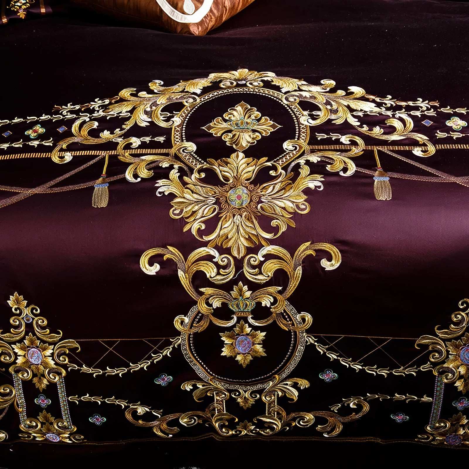 Close-up of maroon bedding showcasing elaborate gold embroidery with baroque-inspired floral and scroll motifs, accented with jewel-like details and intricate tassel designs, emphasizing luxury and opulence.

