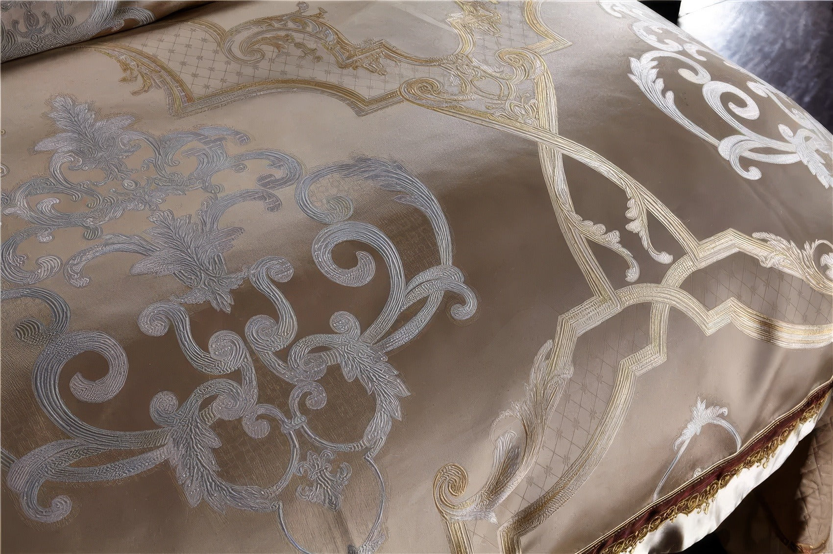 A close-up view of a champagne-toned fabric adorned with intricate baroque-inspired embroidery in pale gold and silver hues. The glossy satin material highlights the opulent details of the swirling floral and geometric patterns, creating a luxurious and sophisticated aesthetic. The interplay of light enhances the depth and richness of the embroidery against the smooth background.

