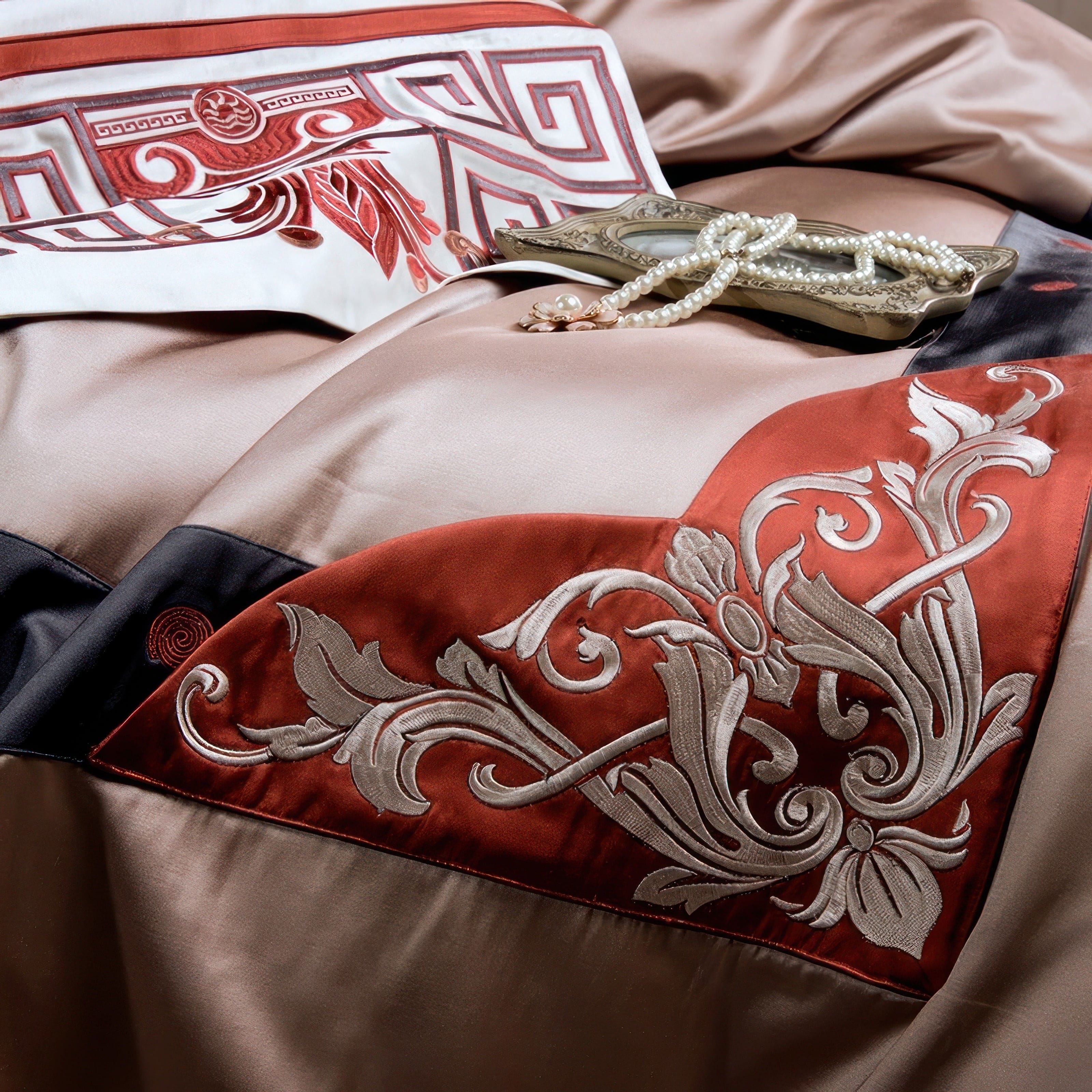 Close-up of a beige fabric featuring intricate red and silver floral embroidery, accented by a bold geometric design in red and white. A vintage-style mirror and pearl necklace with a floral pendant rest on the fabric.