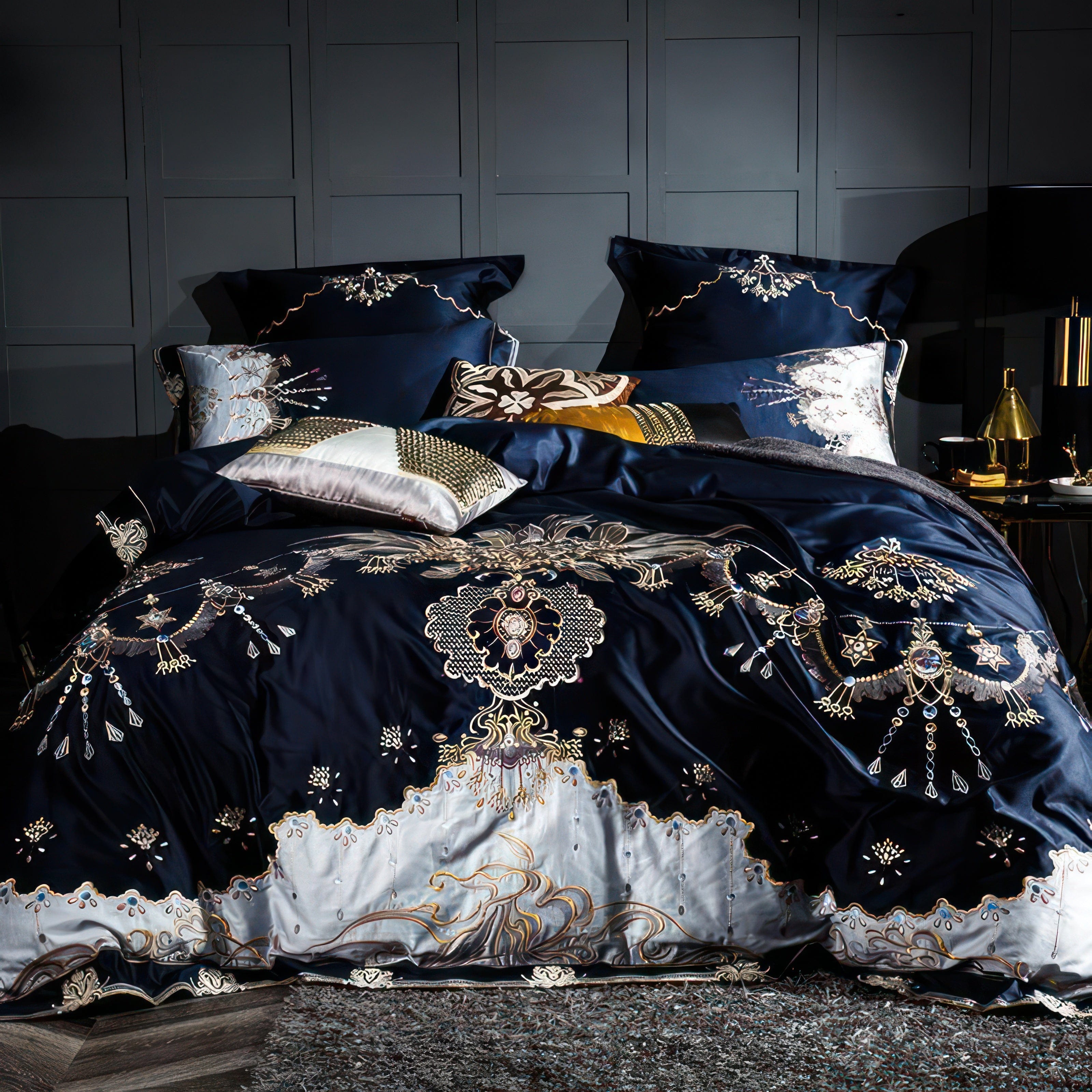 A luxurious bedding set in deep navy with intricate embroidery in gold, silver, and white, featuring floral and ornamental motifs. The design flows seamlessly into a gradient border with silver accents at the base. Decorative pillows in complementary shades of navy, gold, and white enhance the opulent aesthetic. The backdrop includes a modern paneled wall, with a plush gray rug completing the elegant and sophisticated bedroom setting.

