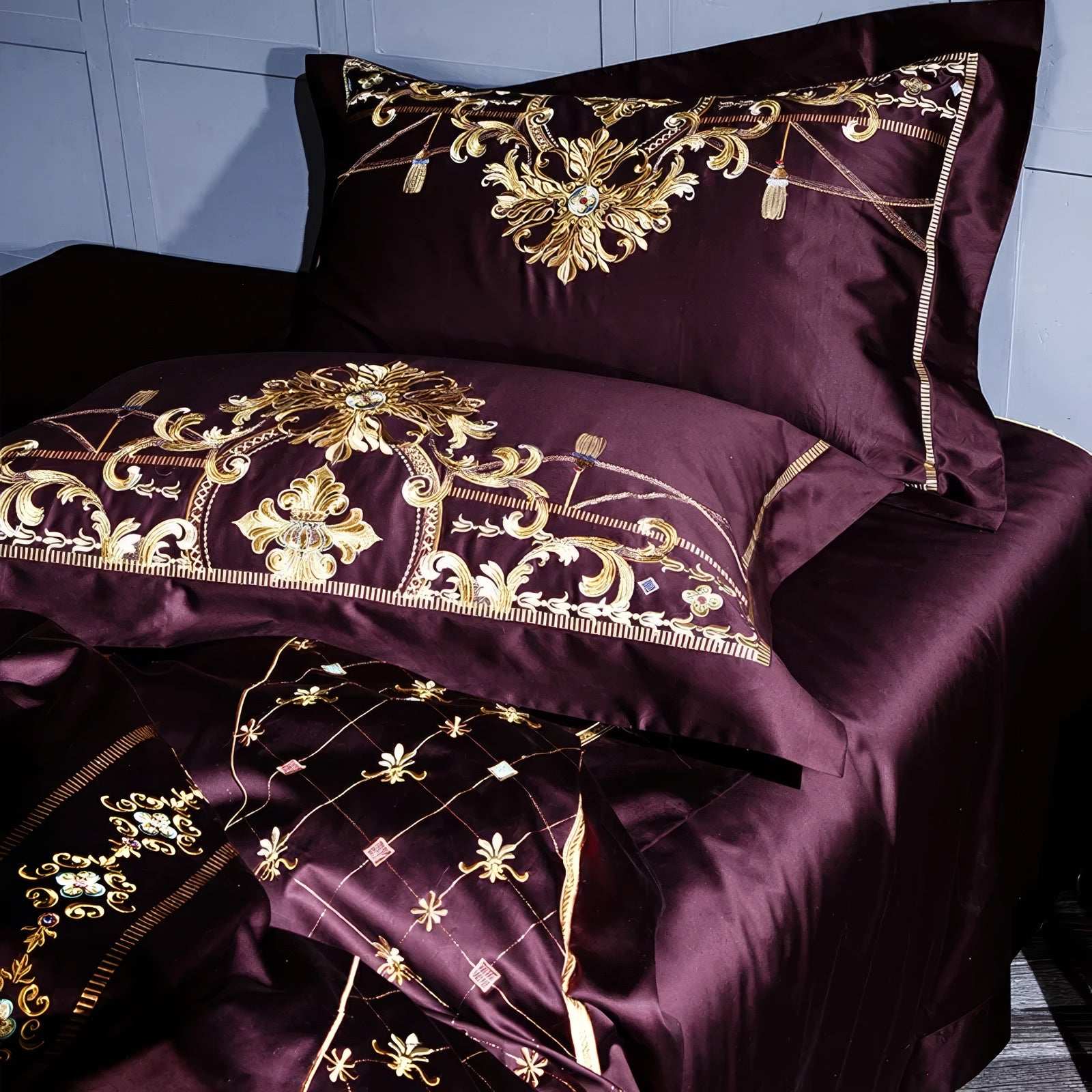 Close-up of maroon bedding featuring intricate gold embroidery with floral and scroll motifs on the pillowcases and sheets, complemented by diamond-patterned accents and fine decorative detailing for an elegant and regal aesthetic.

