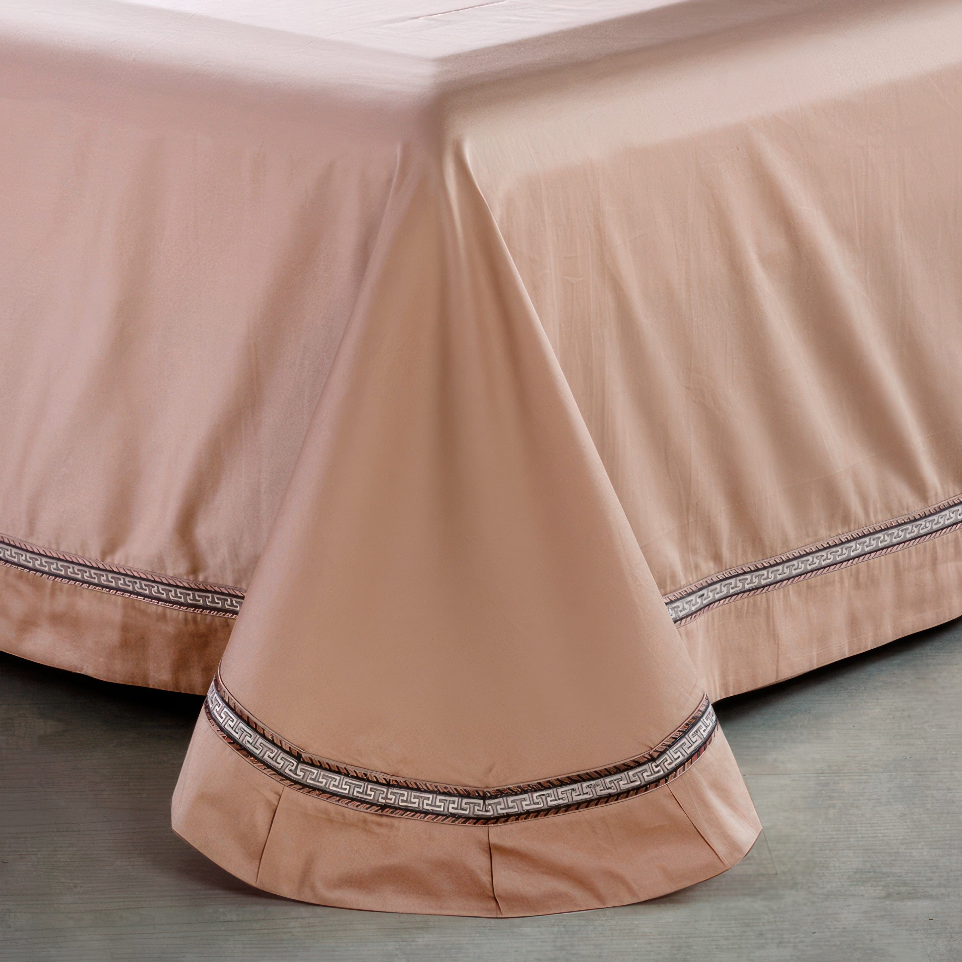 Beige bedskirt with a smooth drape, accented by a decorative trim featuring a metallic Greek key pattern near the hem. The pleated edges add a refined touch, complementing the clean, elegant design.