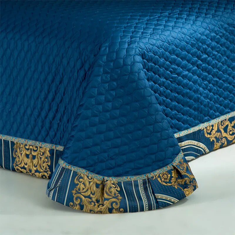 Blue quilted bedspread with a textured diamond pattern, finished with an ornate gold and navy border featuring intricate baroque-style motifs and pleated accents along the edge.

