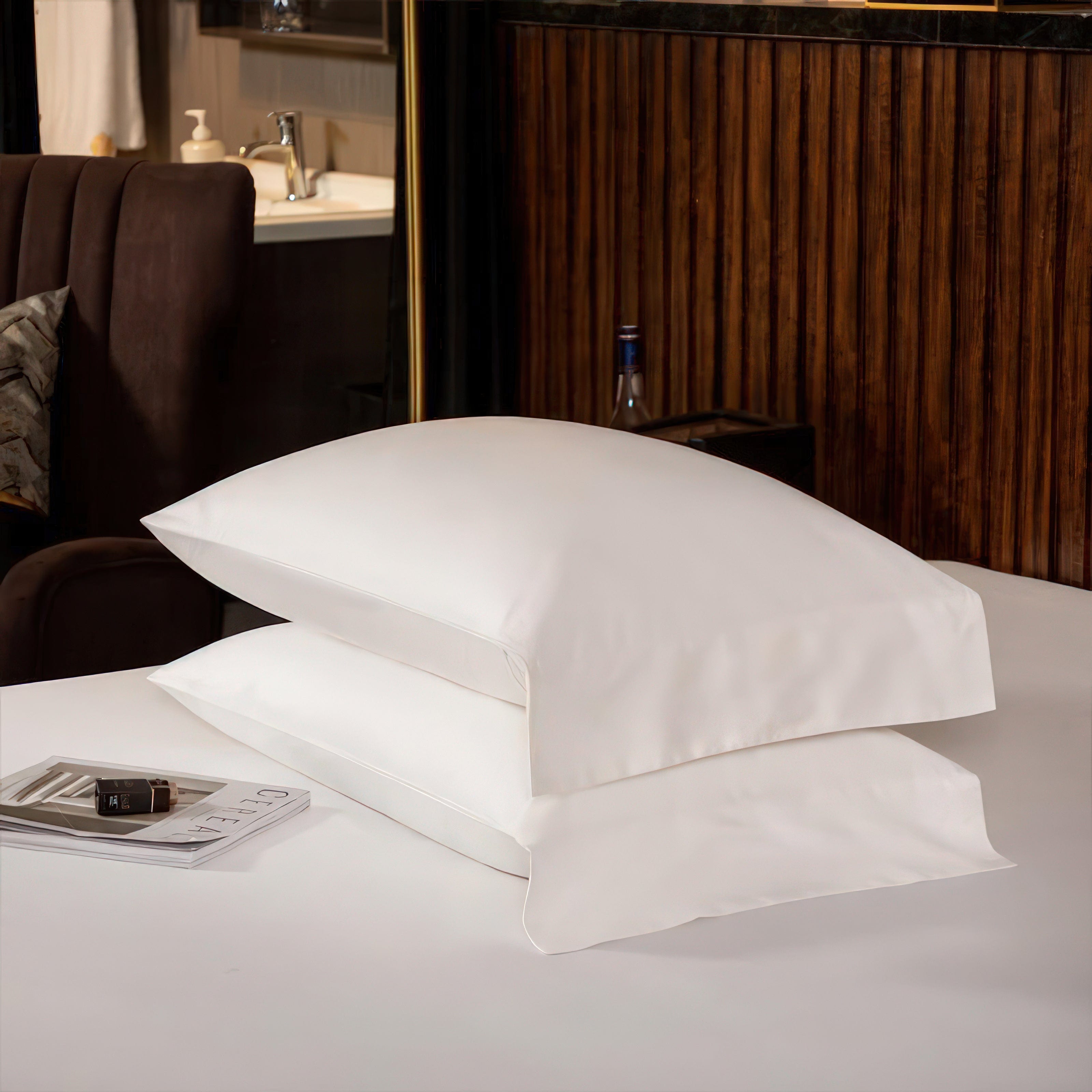 A pair of perfectly stacked white pillows with a smooth and luxurious finish, resting on a white surface. In the background, a contemporary setting with dark wood paneling and a glimpse of a stylish bathroom creates a sophisticated ambiance. A magazine lies nearby, enhancing the minimalistic yet elegant presentation.

