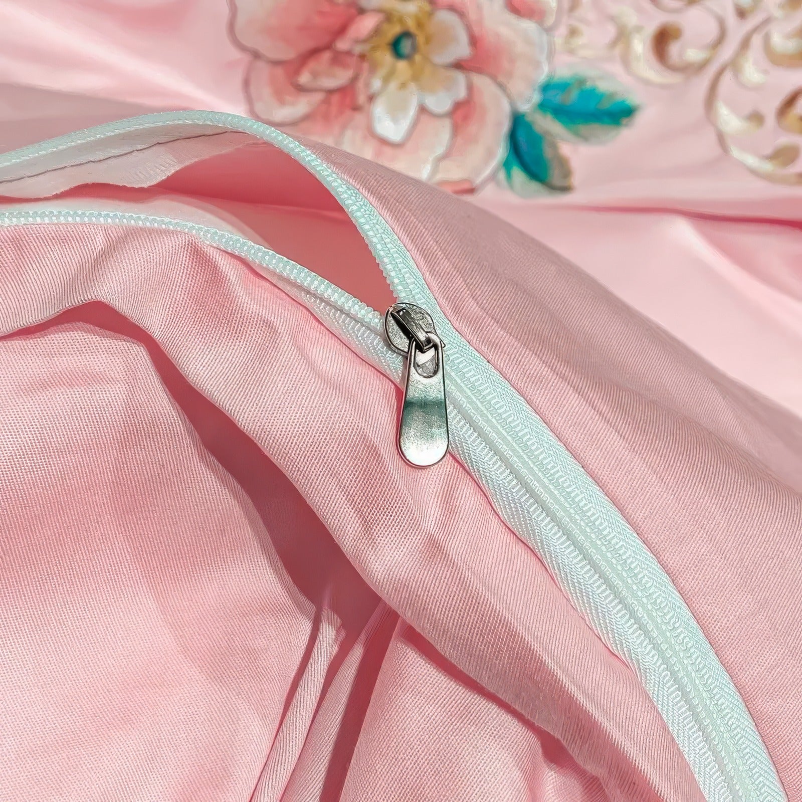 Close-up of a pink duvet cover showcasing a smooth white zipper with a silver pull tab. The detailed view highlights the neat stitching and seamless integration of the zipper into the fabric. In the background, a floral embroidery design in shades of pink, teal, and gold adds a decorative element to the bedding.

