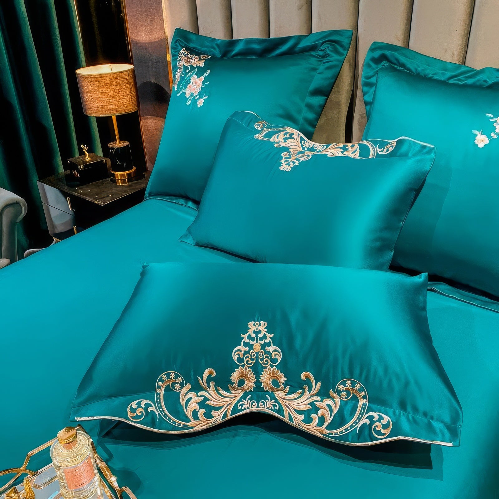 
A close-up of a luxurious teal bedding arrangement featuring a pillow with intricate golden embroidery in a floral motif. The bedding is adorned with matching embroidered details on all pillows, enhancing the regal design. A black side table with a golden lamp and decorative accents complements the opulent setting, adding a warm ambiance.

