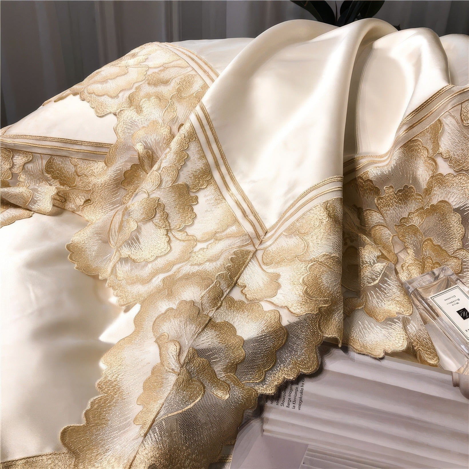 Close-up of white satin bedding with intricate gold lace floral detailing, a folded edge with double stitching, and a perfume bottle resting beside stacked magazines.

