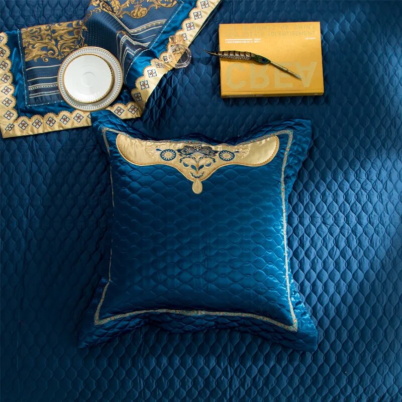 Square quilted navy blue pillow with gold embroidered detailing at the center, placed on a matching quilted bedspread. To the top, a decorative fabric piece with intricate gold designs, a white plate with gold trim, a glass perfume bottle, and a yellow book with a pen complete the arrangement.

