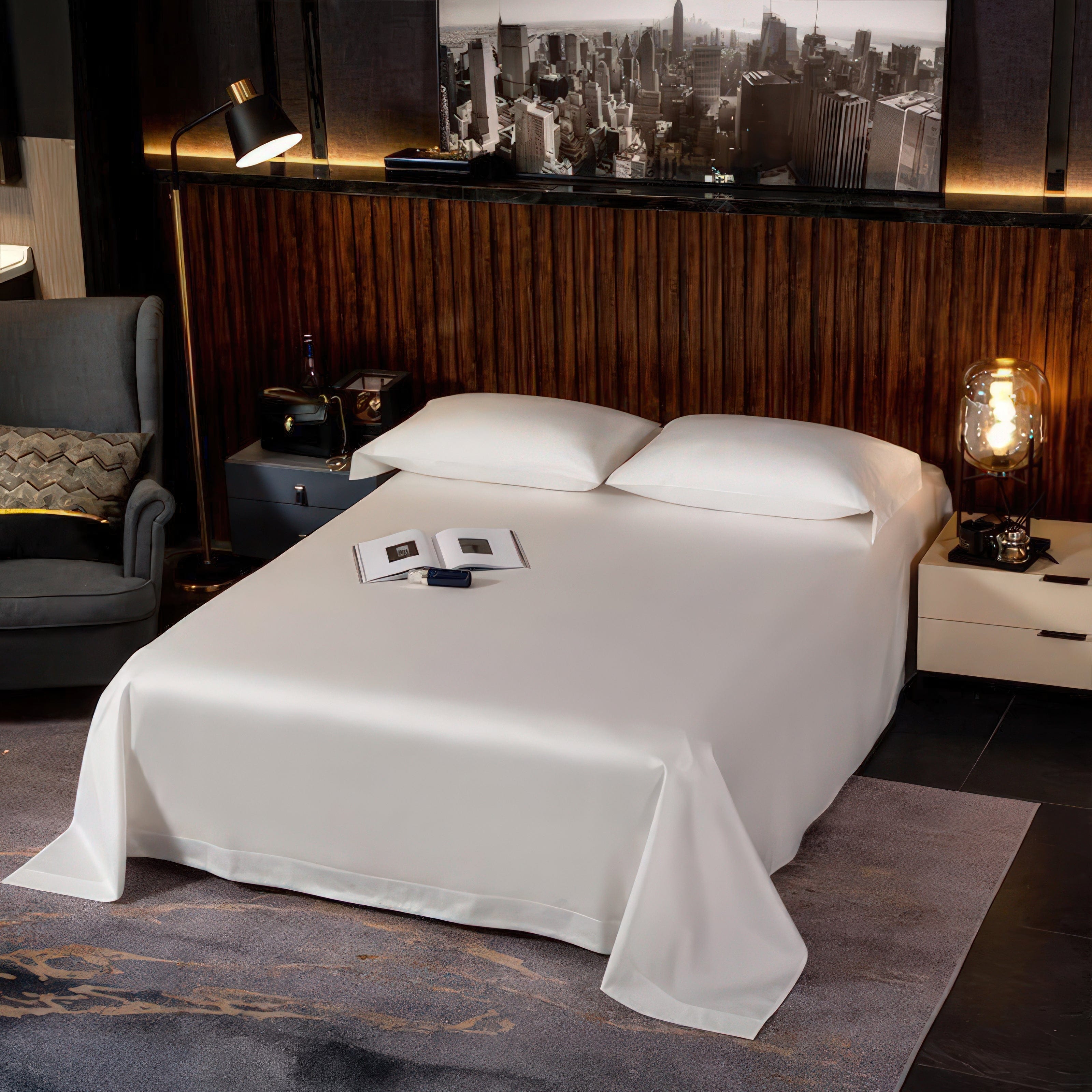 A neatly dressed bed with crisp white bedding, set against a modern and cozy backdrop. The room features warm wooden paneling, a stylish armchair with a textured cushion, and soft ambient lighting. An open book rests on the bed, adding a touch of casual elegance to the minimalist setting. A black-and-white cityscape art piece completes the sophisticated atmosphere.

