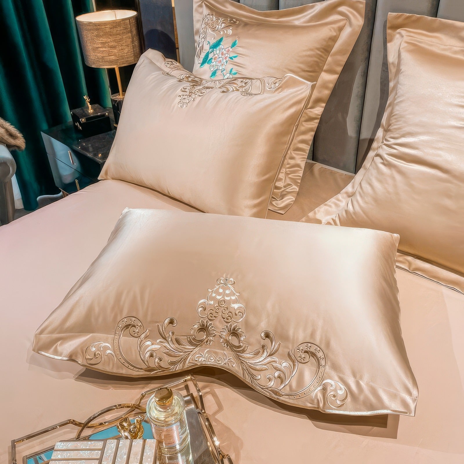 A luxurious golden beige pillowcase adorned with intricate embroidery featuring scroll patterns and a regal floral motif in the center. The pillowcase has a silky sheen that complements its elegant design. Two matching pillows with complementary embroidery and teal floral accents are visible in the background, completing the sophisticated bedding ensemble. A glass tray with decorative items is placed on the bed, enhancing the overall opulent setting.

