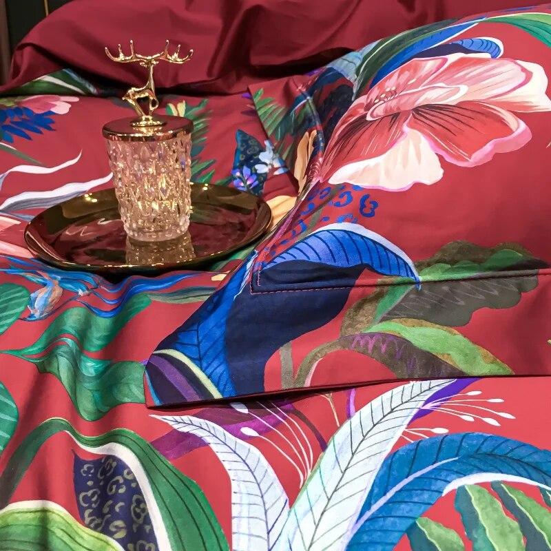 Close-up of a vibrant crimson Egyptian cotton bedding set adorned with tropical floral and botanical patterns in vivid hues of blue, green, and pink. A crystal jar with a golden lid rests on a polished gold tray, adding a touch of elegance and luxury to the setting.

