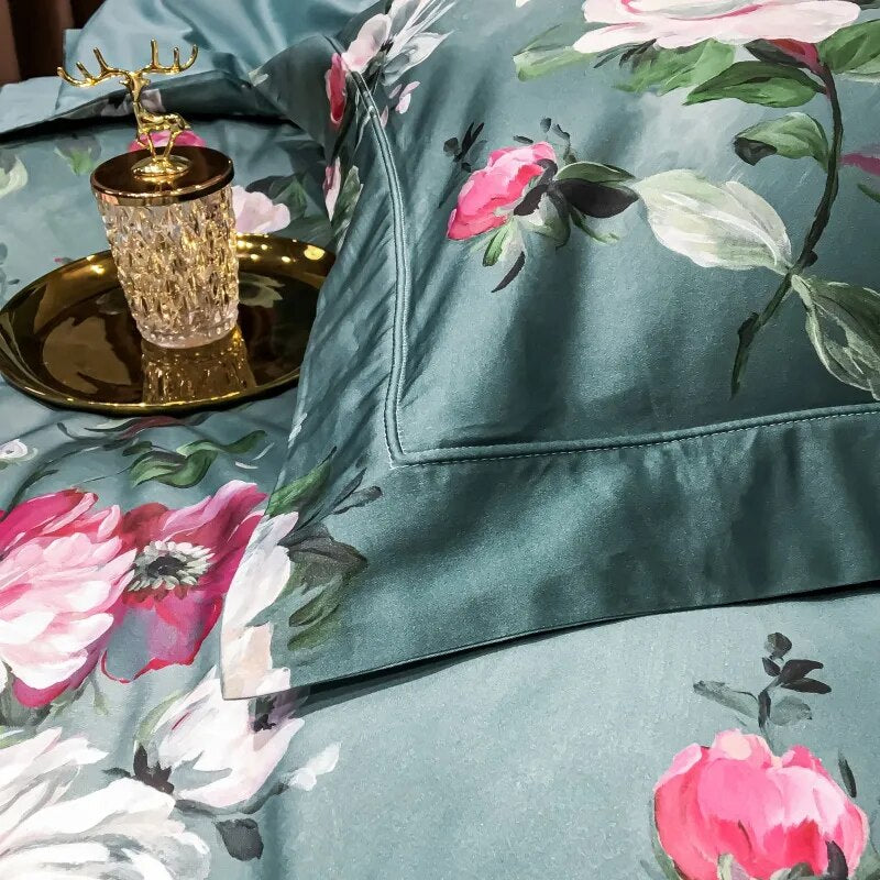 Close-up of a pillowcase from an Egyptian cotton bedding set, featuring a smooth teal fabric adorned with vibrant pink and white floral patterns. The detailed stitching emphasizes the luxurious quality, complemented by a golden tray with a crystal glass and deer ornament for an elegant touch.

