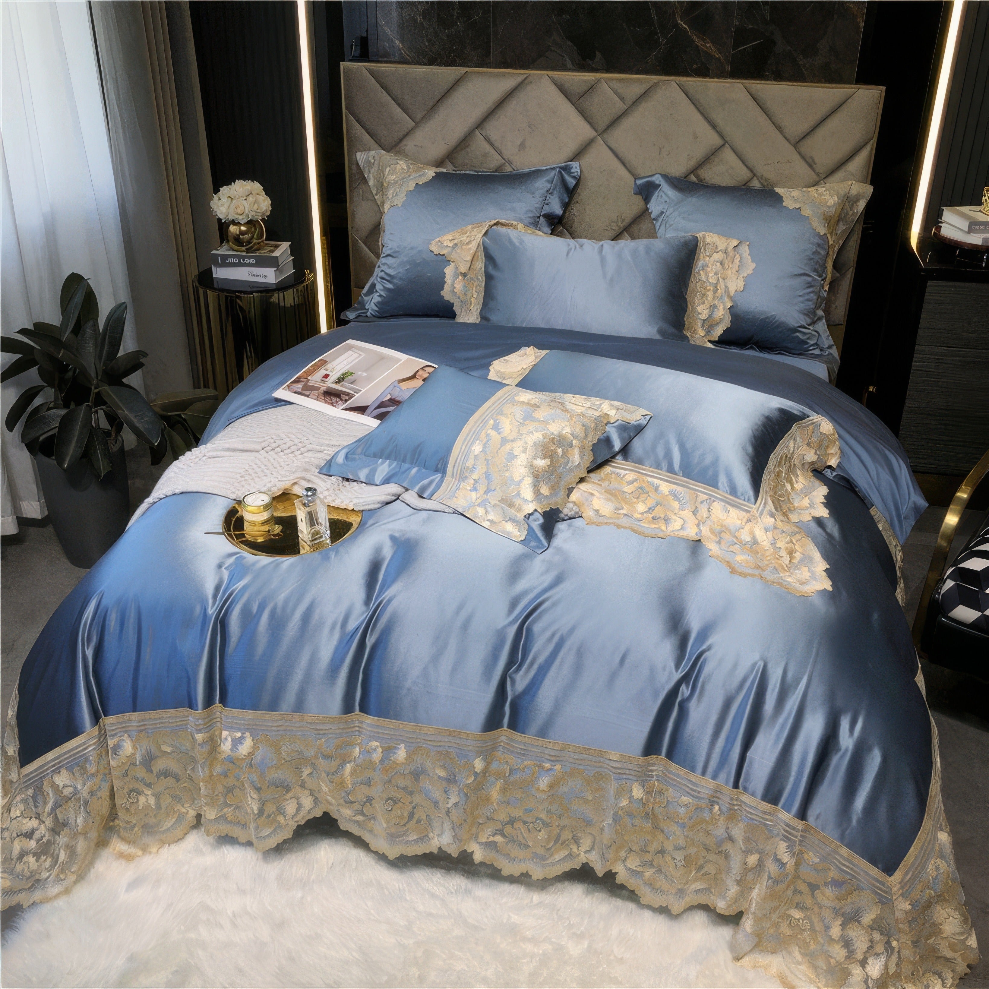 Elegant blue satin bedding set with intricate gold lace detailing, accompanied by decorative pillows and a neatly placed tray holding candles and perfume atop a soft white throw blanket.

