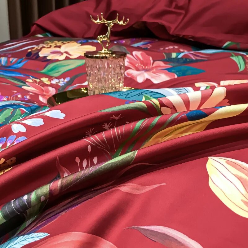 Close-up of a crimson Egyptian cotton duvet showcasing vibrant tropical floral prints in bold shades of pink, yellow, green, and blue. The silky smooth texture highlights the intricate botanical details, with a golden decorative tray and crystal accent adding elegance to the luxurious bedding ensemble.

