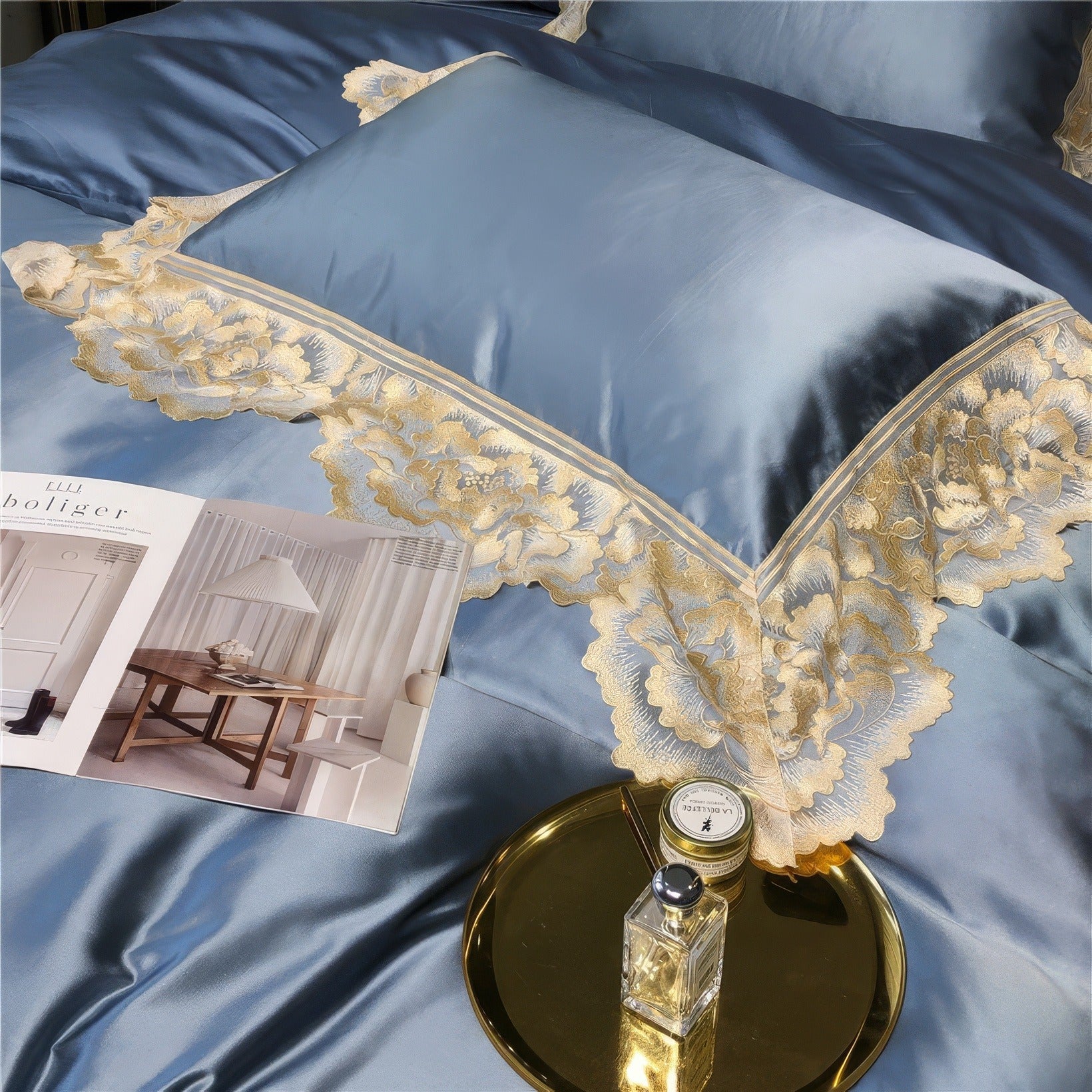 Close-up of blue satin bedding with gold lace floral embroidery, a gold tray holding a glass perfume bottle, and a small candle, alongside an open interior design magazine.

