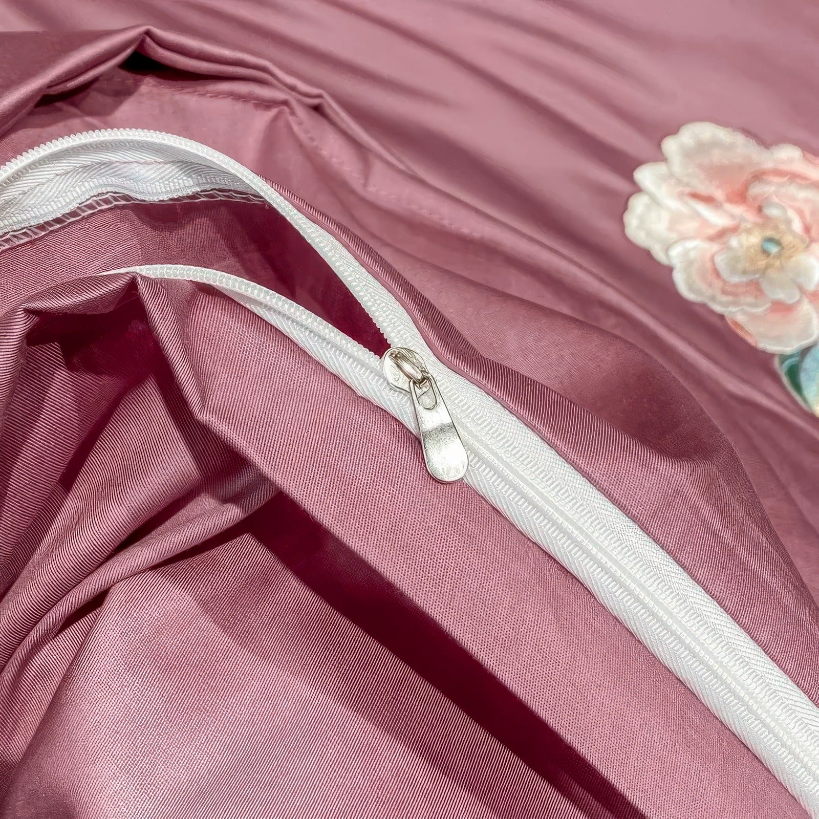 A detailed close-up of a mauve silk duvet cover showcasing a high-quality white zipper closure, designed for ease of use and secure bedding encasement. The soft fabric texture and floral embroidery in the background add a touch of elegance and luxury.

