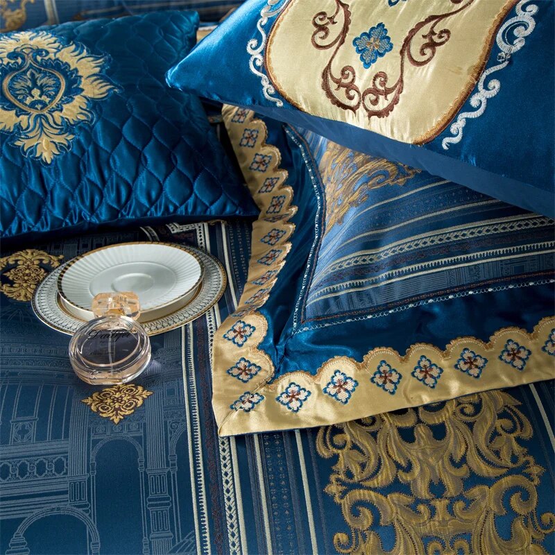 Luxurious bedding with intricate blue and gold embroidery features a mix of quilted and satin textures. A matching pillowcase with ornate gold detailing complements the fabric's design. A white plate with a gold rim and a clear glass perfume bottle rest on the bed, adding a touch of opulence to the scene.

