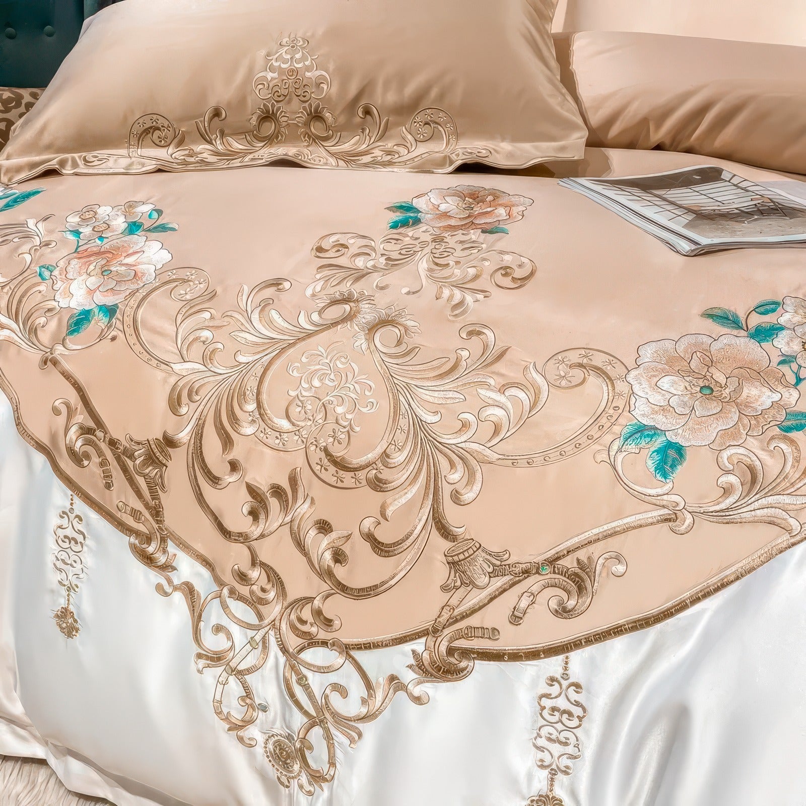 A close-up of a luxurious golden beige duvet cover showcasing intricate embroidery. The design features elegant floral patterns with gold accents and teal highlights, surrounded by delicate scroll motifs. The embroidery cascades seamlessly onto a white satin base, enhancing the bedding's opulent appeal. A matching pillowcase with complementary embroidery is partially visible, adding cohesion to the sophisticated design.

