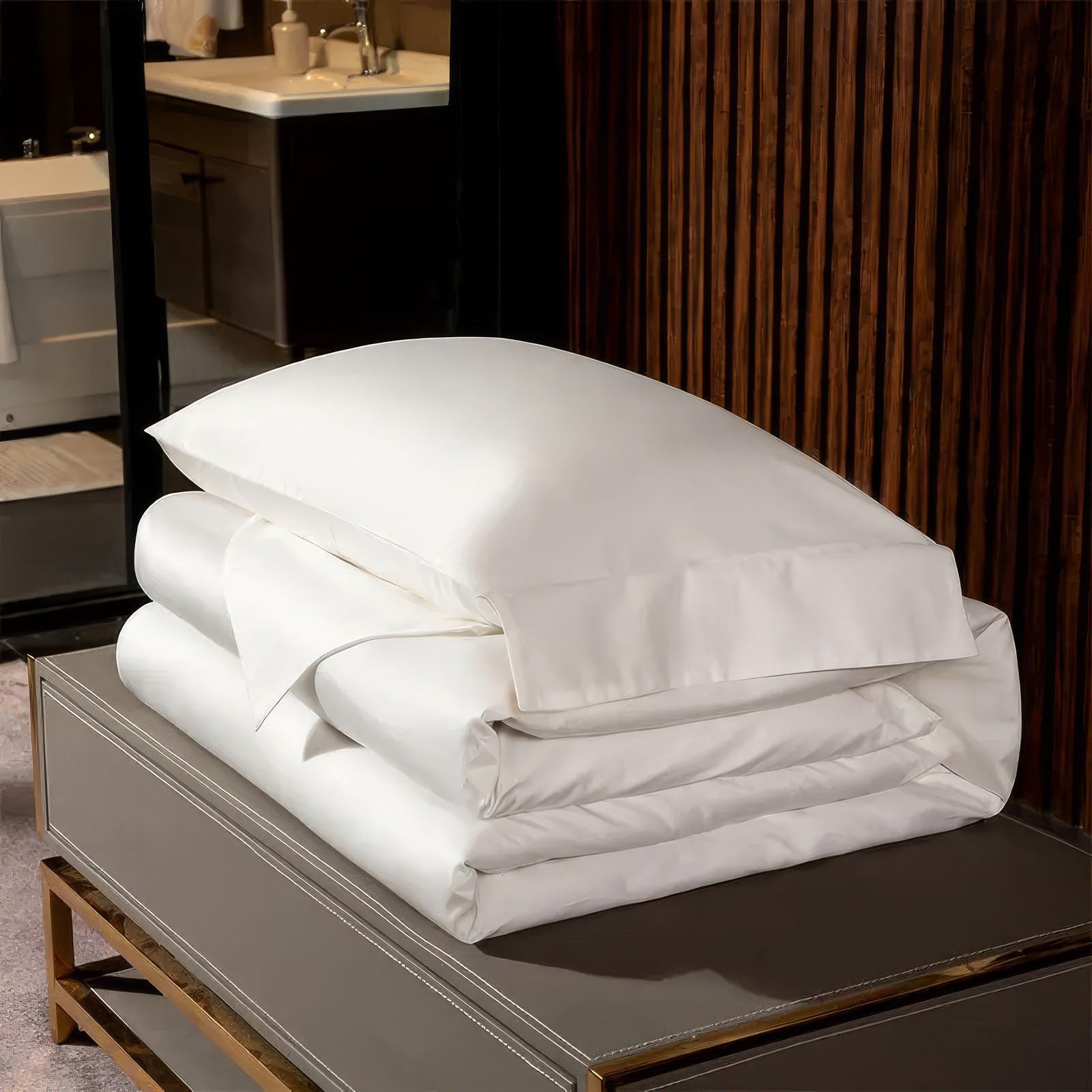 Stacked white bedding set, including a pillowcase, duvet, and sheets, neatly folded on a beige bench against a backdrop of dark wooden panels and a partial view of a bathroom.

