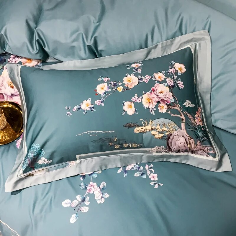 Teal pillowcase featuring an intricate floral design with blooming cherry blossoms in soft pink, white, and yellow. The detailed scene includes a tree, rocks, and delicate foliage, creating an elegant, artistic landscape. The pillowcase has a smooth sheen and subtle border trim, matching the luxurious teal bedding beneath it. A golden tray with a crystal ornament is partially visible nearby.

