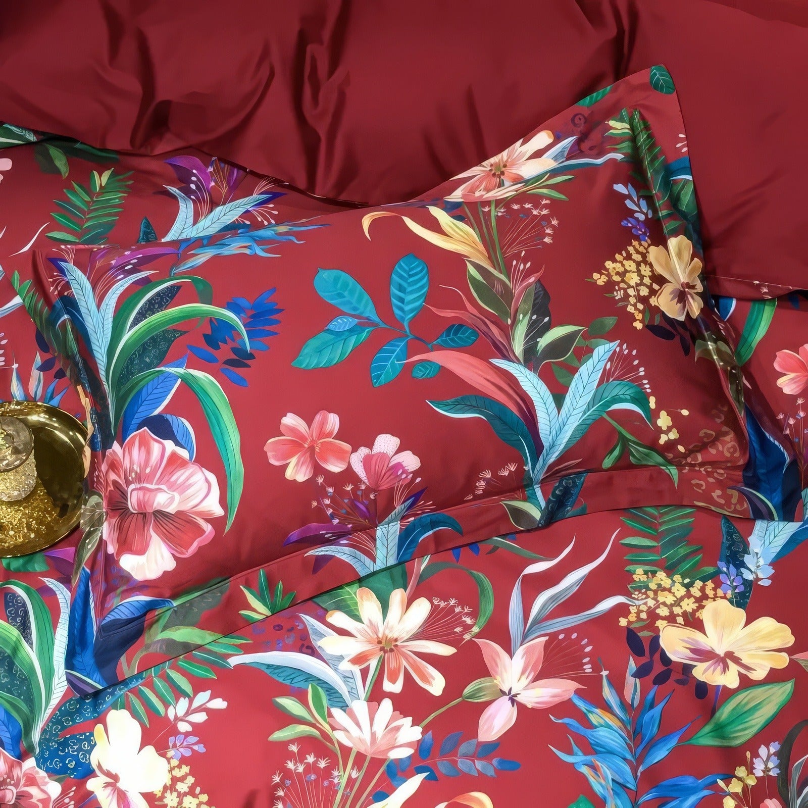 Vibrant crimson Egyptian cotton pillowcases adorned with tropical floral prints in a lively mix of pink, yellow, blue, and green. The smooth fabric highlights intricate patterns of exotic leaves and blossoms, complemented by a matching red duvet cover for a bold and luxurious look. A golden tray with decorative accents adds a touch of sophistication to the setting.

