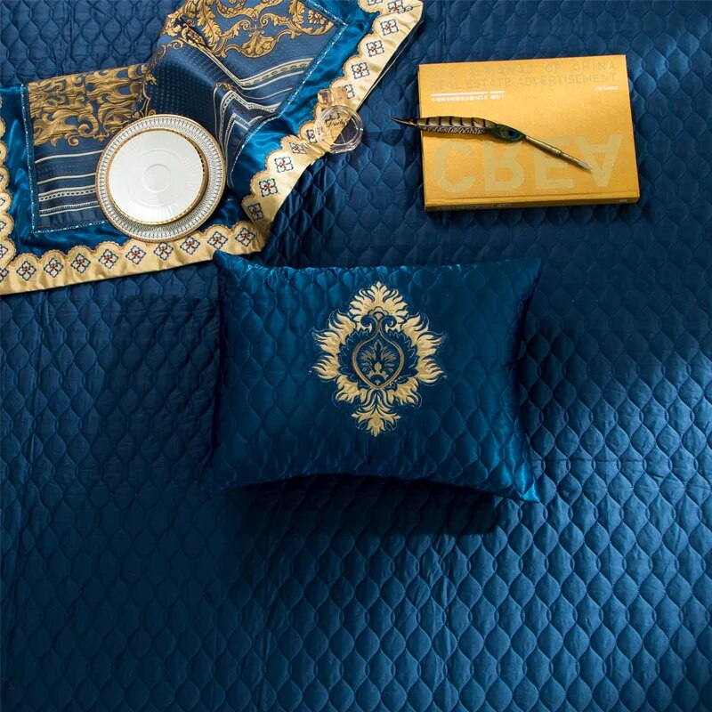 Quilted navy blue bedding with a decorative pillow featuring a gold embroidered emblem in the center. A matching ornate fabric piece with gold detailing, a white plate with gold trim, a glass perfume bottle, and a yellow book with a pen rest on the bed, complementing the luxurious aesthetic.

