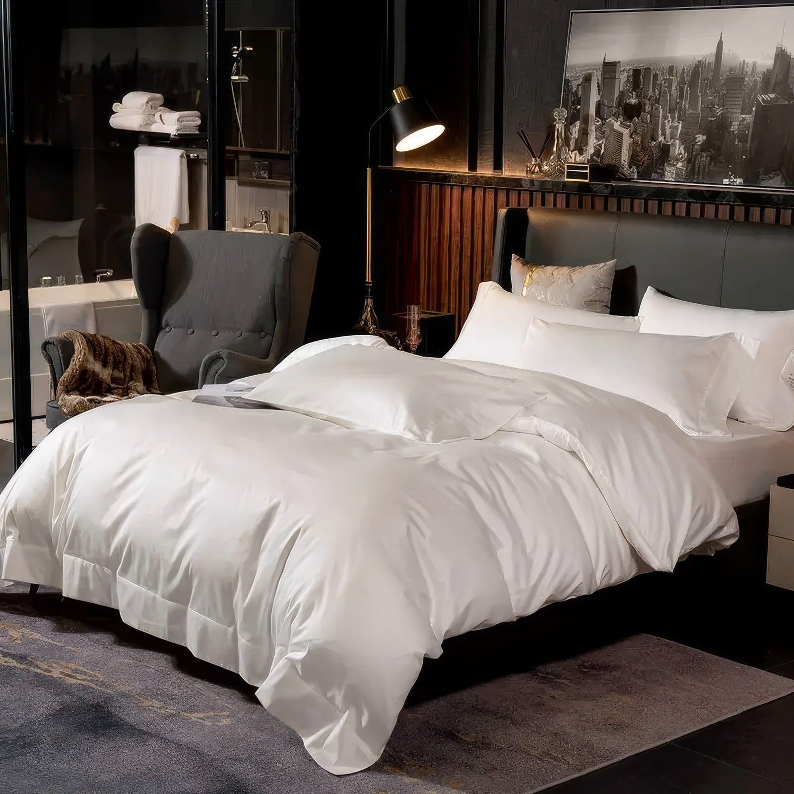 White bedding set in a luxurious, modern bedroom with a dark wood headboard, a black and white cityscape photograph, and a gray armchair near a bathroom with stacked towels.

