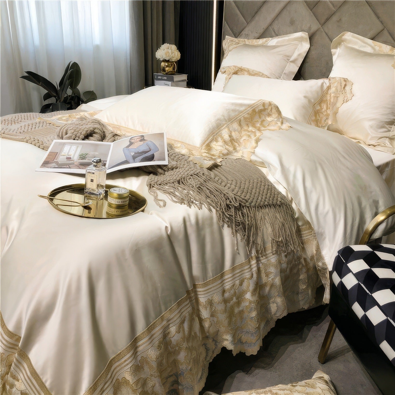 Luxurious white satin bedding with gold lace detailing, a beige knit throw, a gold tray holding a perfume bottle and cream jar, and a magazine on the bed.

