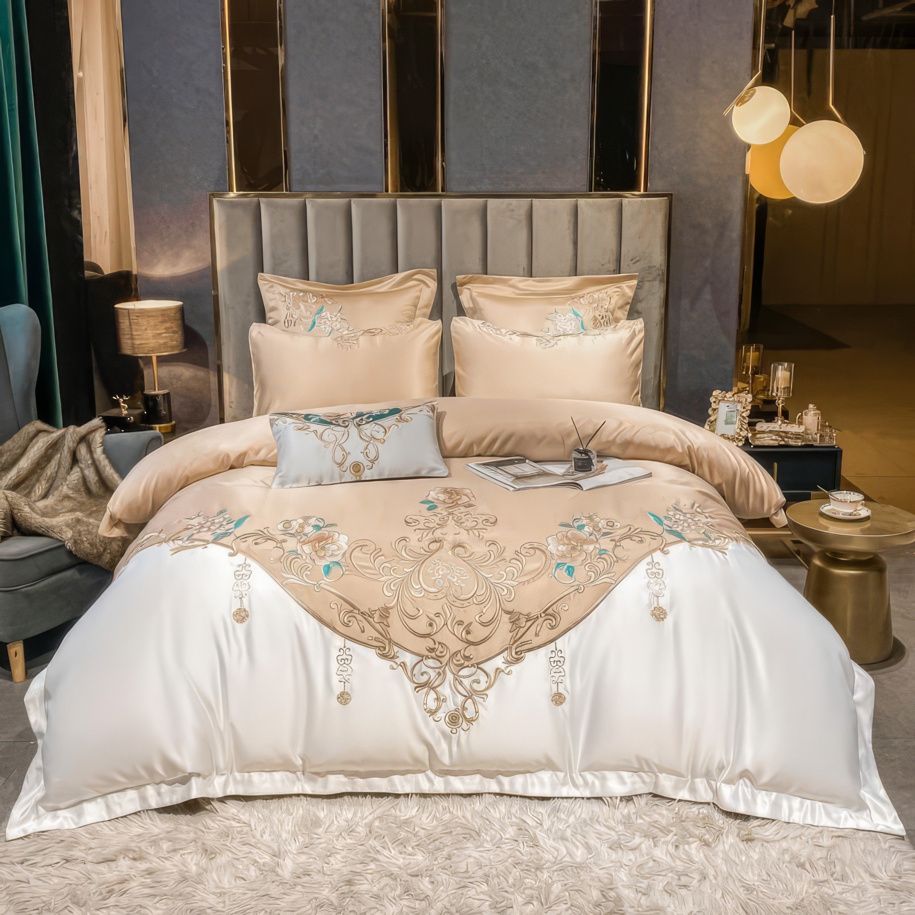 A luxurious bedding set featuring a golden beige duvet cover with intricate floral and scroll embroidery in soft gold and teal hues. The design cascades elegantly onto a white satin base, creating a striking contrast. Complementary pillowcases with matching embroidery are layered against a tufted gray headboard, adding depth and texture. The bedroom is styled with a blend of modern and classic accents.