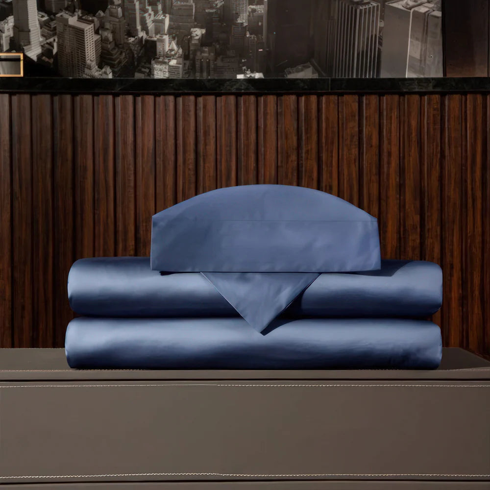 Folded blue satin bedding set displayed on a gray surface, consisting of pillowcases, a flat sheet, and a fitted sheet, with a dark wood-paneled wall and a cityscape photo in the background.

