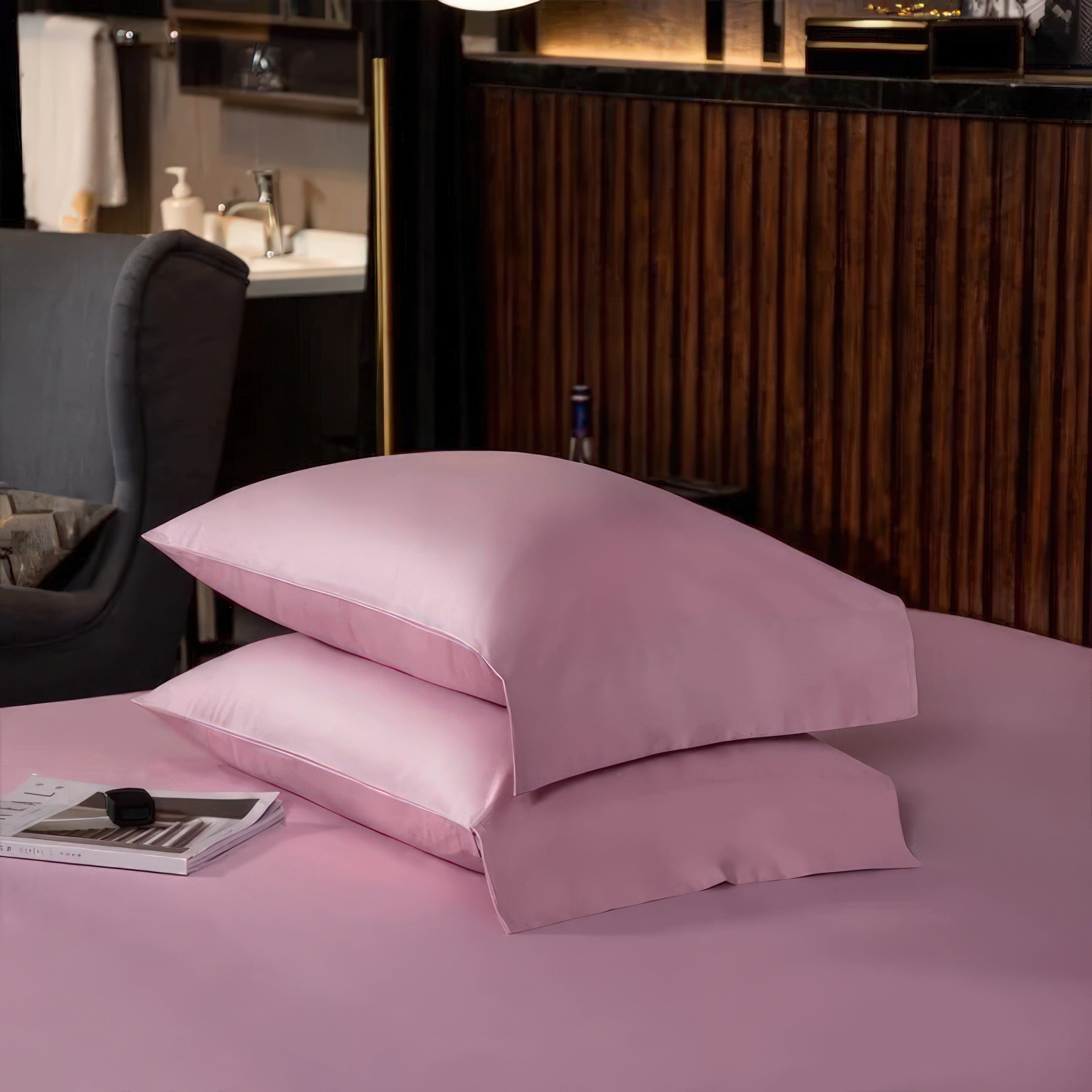 Two neatly stacked soft pink pillows placed on a matching pink bedspread, set in a modern bedroom with a cozy ambiance. The background features a stylish armchair, a wooden-paneled wall, and glimpses of a bathroom area, creating a balance between elegance and functionality. A magazine and a sleek smartwatch on the bed add subtle details of relaxation and modern living.

