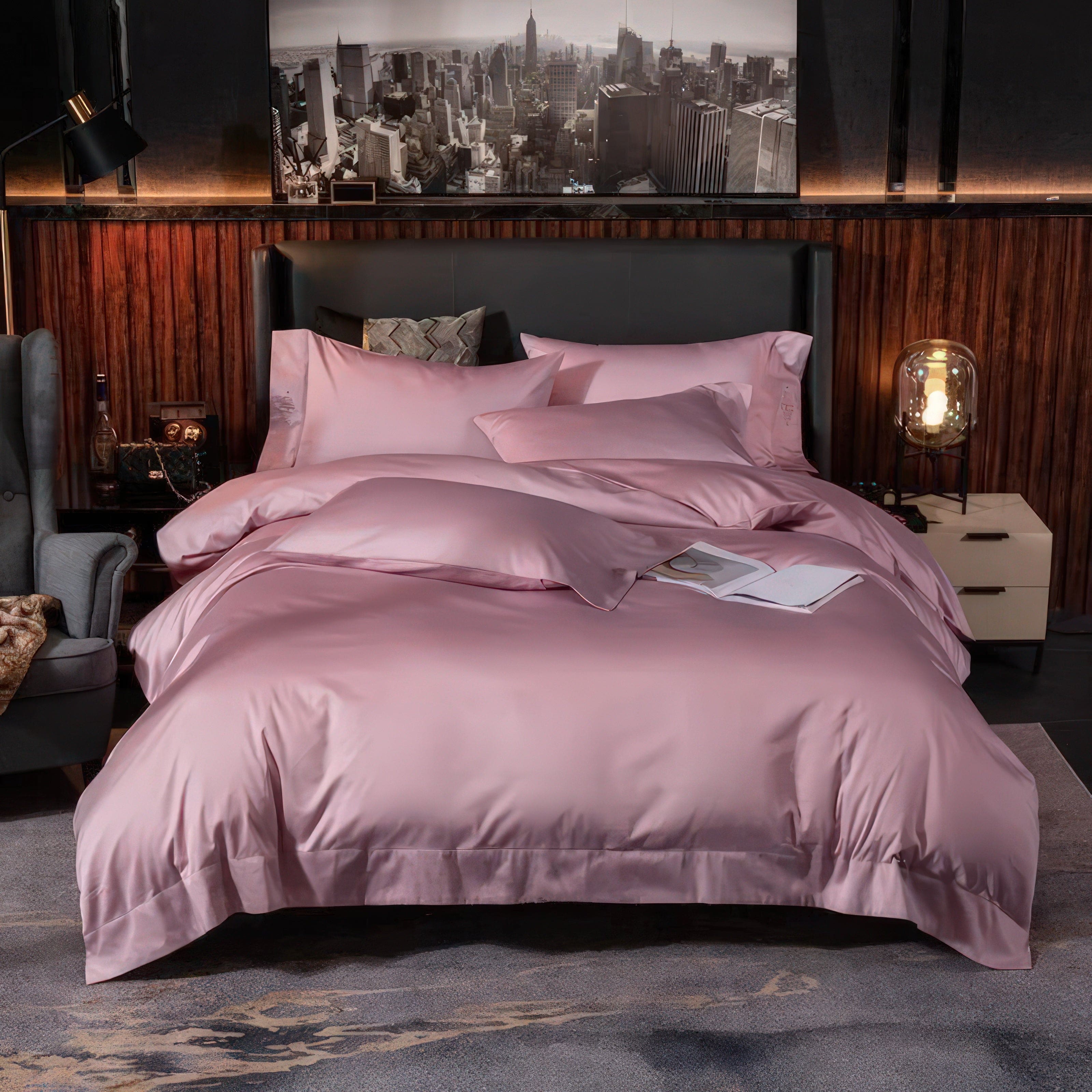 A luxurious bed adorned with soft pink bedding, radiating elegance and warmth. The setting is modern with a wooden-paneled backdrop, complemented by a sophisticated armchair and ambient lighting. The decor includes a black-and-white cityscape artwork above the headboard, adding a cosmopolitan touch. A book casually placed on the bed enhances the cozy and inviting atmosphere.

