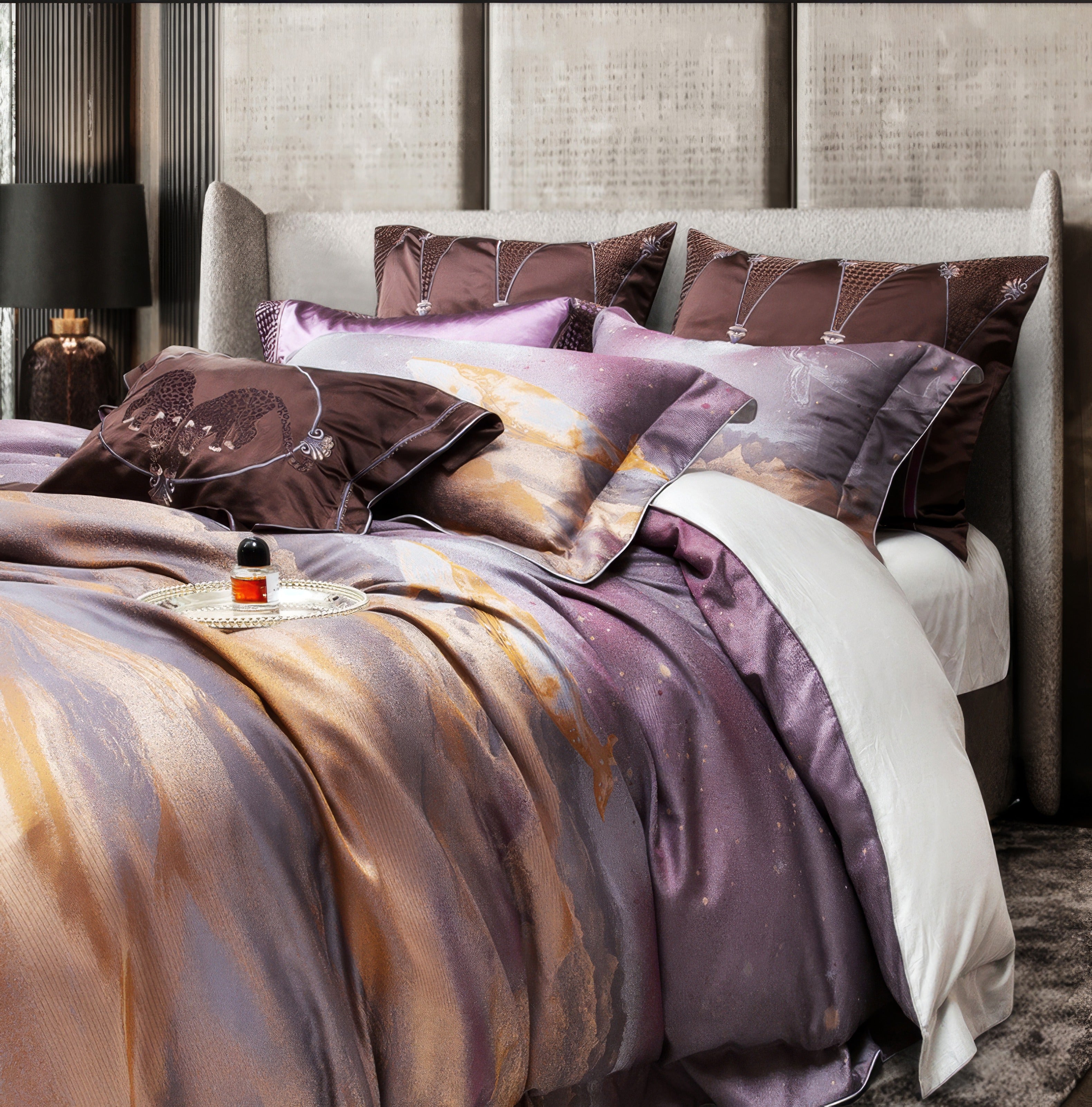 A close-up view of a luxurious bedding set showcasing a harmonious blend of purple, gold, and grey tones in an abstract artistic design. The bedding includes matching pillowcases with sleek piping and embroidery details, complemented by decorative accent pillows in rich brown tones featuring intricate patterns. The setup exudes elegance with its plush fabrics and layered textures, set against a modern light-grey upholstered headboard. A silver tray with a small perfume bottle adds a touch of opulence, enhan