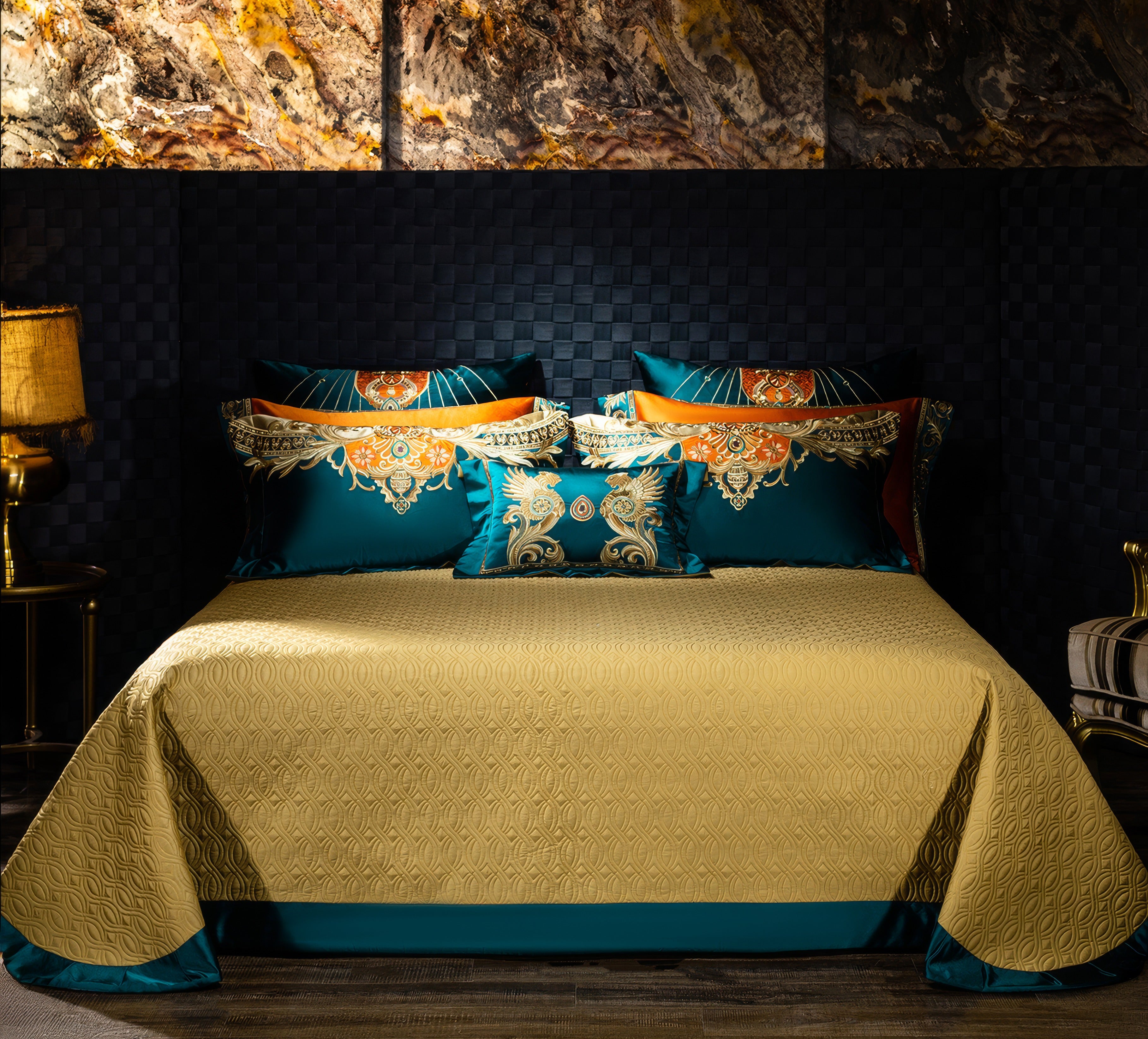 Luxurious bedding set featuring a golden quilted bedspread with teal and orange accents, complemented by pillowcases and a decorative throw pillow with intricate gold and turquoise baroque-style embroidery, styled in a sophisticated bedroom with a textured black wall and marble accents.