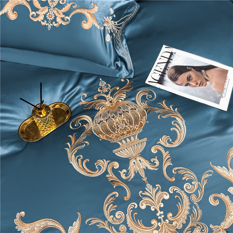 Blue satin bedding with intricate gold embroidery, featuring a centerpiece urn surrounded by ornate floral patterns. A gold tray with a diffuser sits beside a fashion magazine on the bed.

