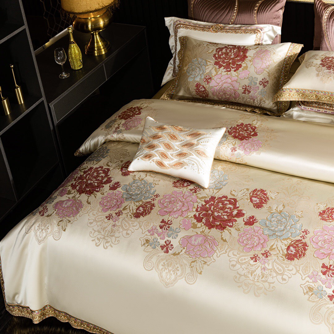 A close-up view of a luxurious bedding set showcasing an ivory duvet cover with elaborate floral patterns in red, pink, and light blue hues. The design features golden accents and intricate embroidery along the edges. A decorative square pillow with an elegant golden wave pattern complements the set. The setup includes matching floral pillowcases and quilted mauve pillows in the background, highlighting the richness and sophistication of the design. The bedside features a golden lamp and accessories, enhanc