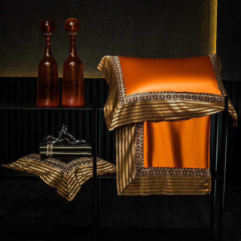 Elegant orange satin bedding with ornate gold and black embroidered trim, displayed folded over a rack. Next to it are two amber glass vases and a decorative black and gold sculpture, creating a luxurious and sophisticated setting.

