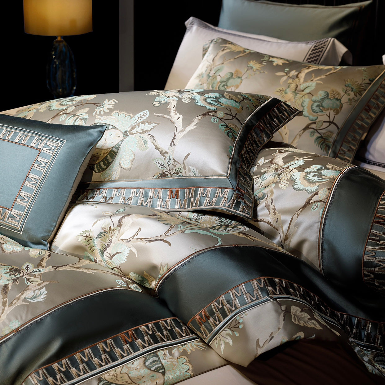 This close-up image captures the luxurious details of a premium bedding set featuring intricate floral and butterfly patterns in soft beige and sage green hues. The fabric showcases a silky texture, enhanced by elegant embroidery and geometric trim accents. The layered pillows display a harmonious blend of designs, including a rectangular decorative pillow with a bordered frame motif. The lighting highlights the glossy finish and fine craftsmanship, while a glowing lamp in the background adds a touch of war