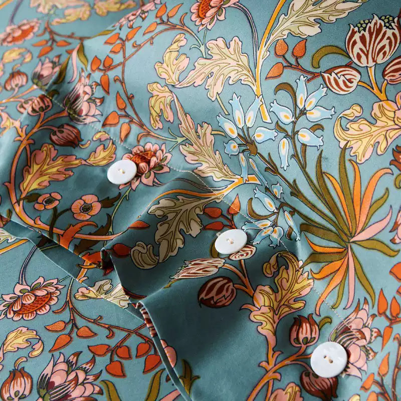 Close-up of a teal pillowcase featuring intricate botanical patterns with orange, red, and pale blue flowers, green leaves, and detailed vines. The white buttons and neat stitching highlight the functional closure of the pillowcase.

