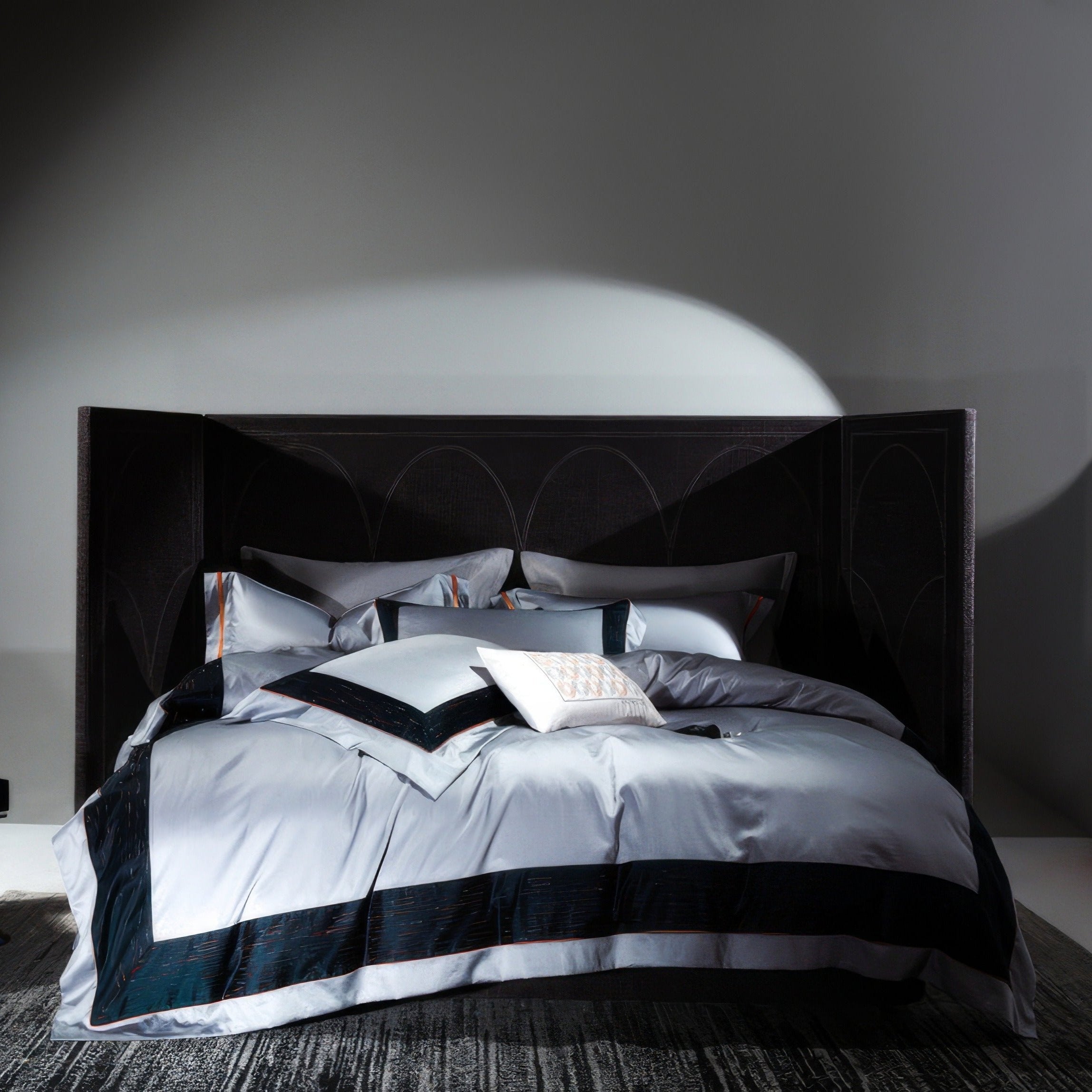 Luxurious bedroom featuring a contemporary bed with an oversized dark headboard and elegant bedding in shades of silver and deep navy blue. The bedding includes a duvet cover with bold navy borders, neatly folded pillows, and a decorative white cushion with intricate patterns. Soft lighting highlights the smooth, high-quality fabric, creating a modern and sophisticated ambiance. The room has a dark carpet, enhancing the dramatic yet calming atmosphere.

