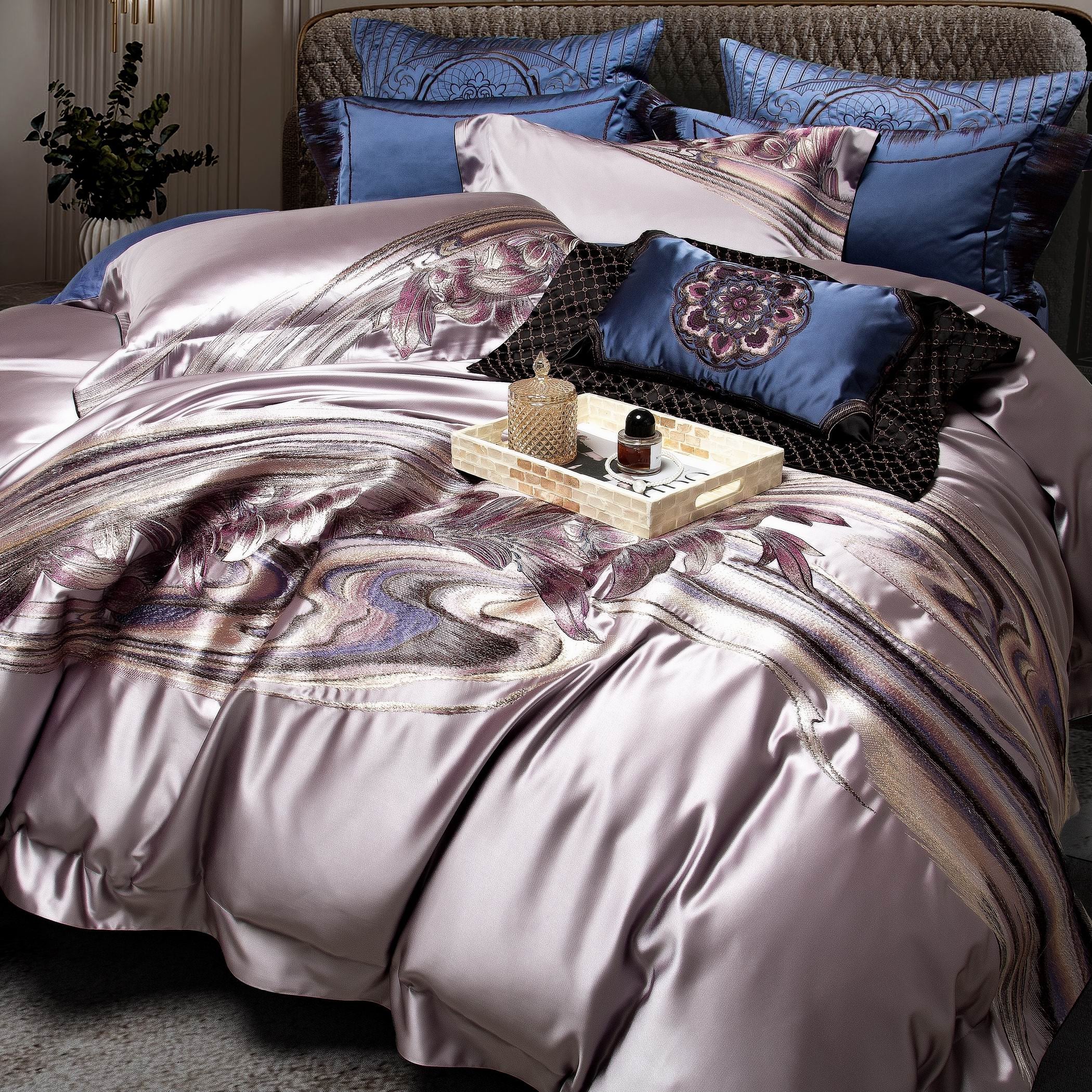 A lavender-gray duvet with intricate floral and swirl embroidery in purples and earthy tones, paired with matching blue pillowcases featuring detailed stitching. The centerpiece is a navy blue decorative cushion with floral embroidery and black textured accents. A mosaic tray on the bed holds a gold-textured jar and a small perfume bottle, adding a touch of elegance. The setting is completed with a quilted headboard and subtle greenery in the background.