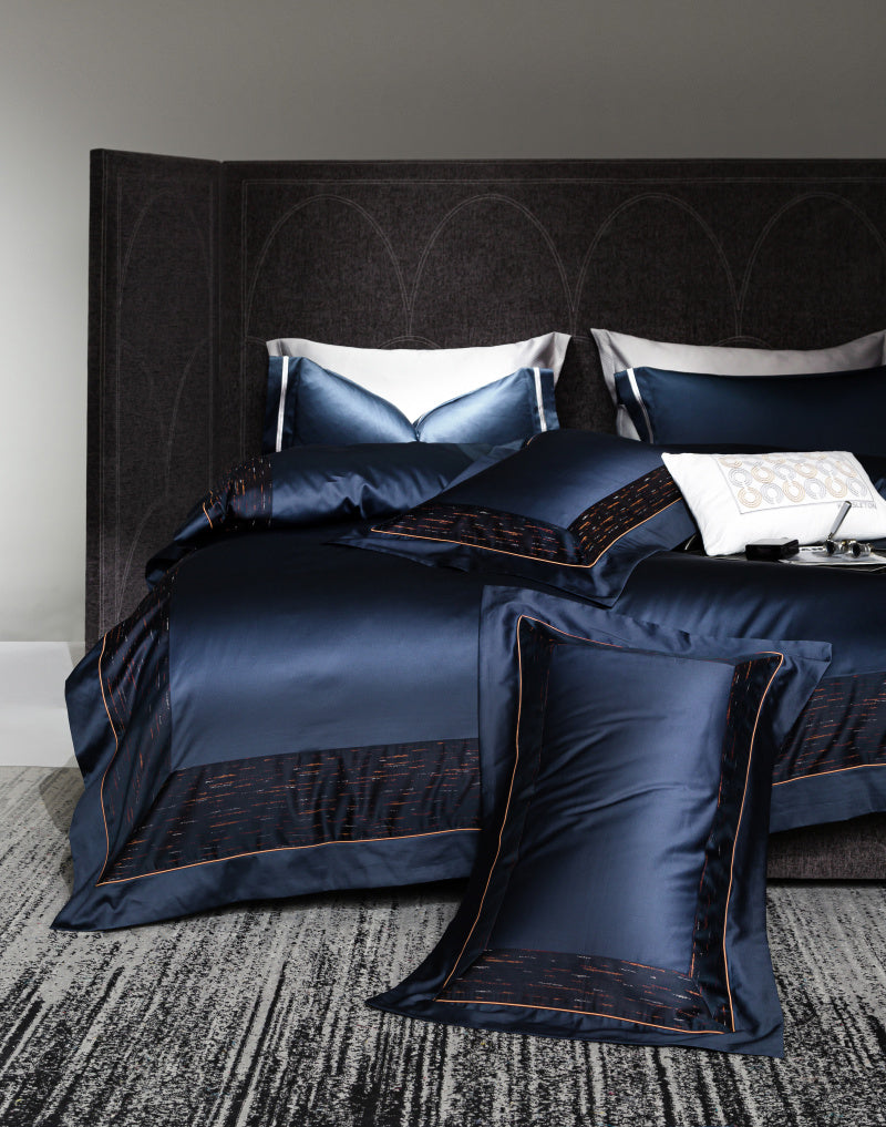 Dark navy blue luxury bedding set with black textured borders and subtle red stitching. The bedding includes matching pillowcases, a decorative white cushion with embroidered details, and a large accent pillow on the floor. The bed is against a dark arched-panel headboard, enhancing the modern and sophisticated design. A textured grey rug lies beneath the bed, completing the contemporary bedroom ambiance.

